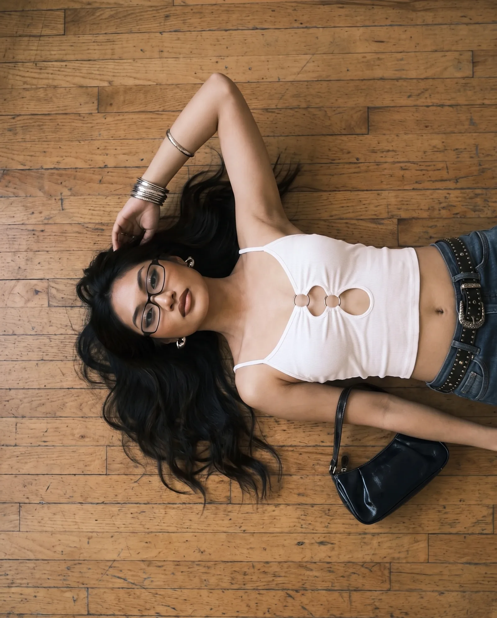 Overhead shot of the white ring-cutout cami and studded-belt jeans, lying on a bare wood floor beside a black shoulder bag.