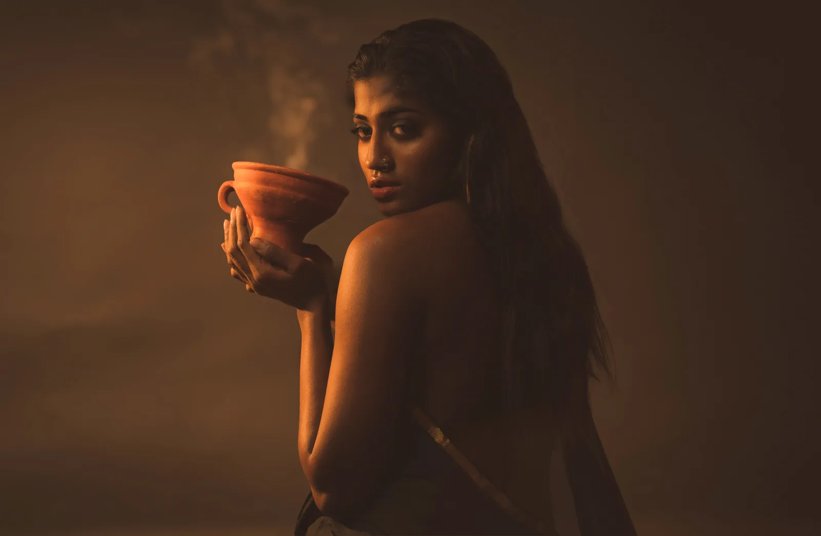 Woman holding a smoking clay incense pot close to her face, bare back to the camera, long hair falling down her back, looking back over her shoulder.