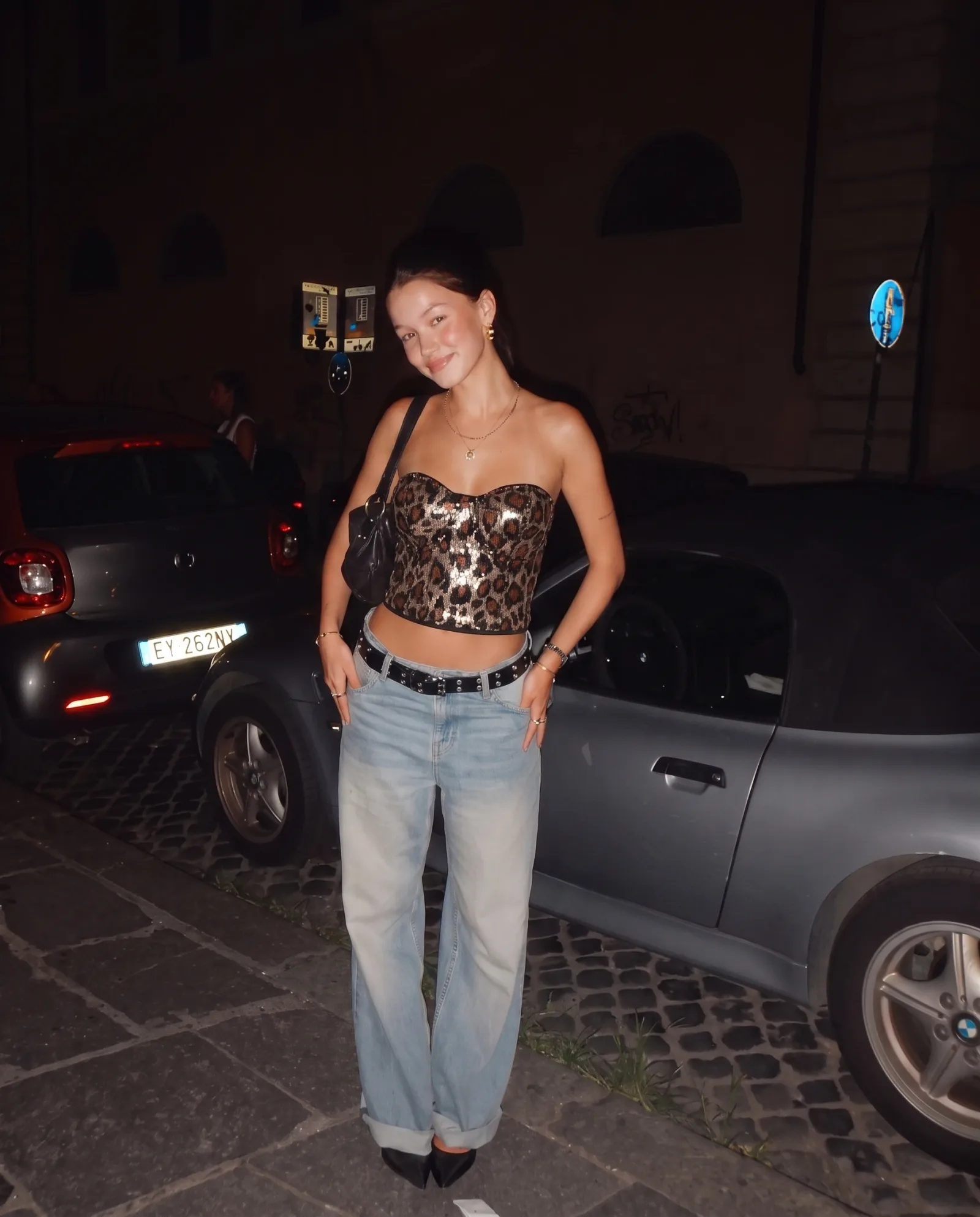 Leopard sequin strapless bustier with jeans on a night street, candid smile, shopfront light behind.