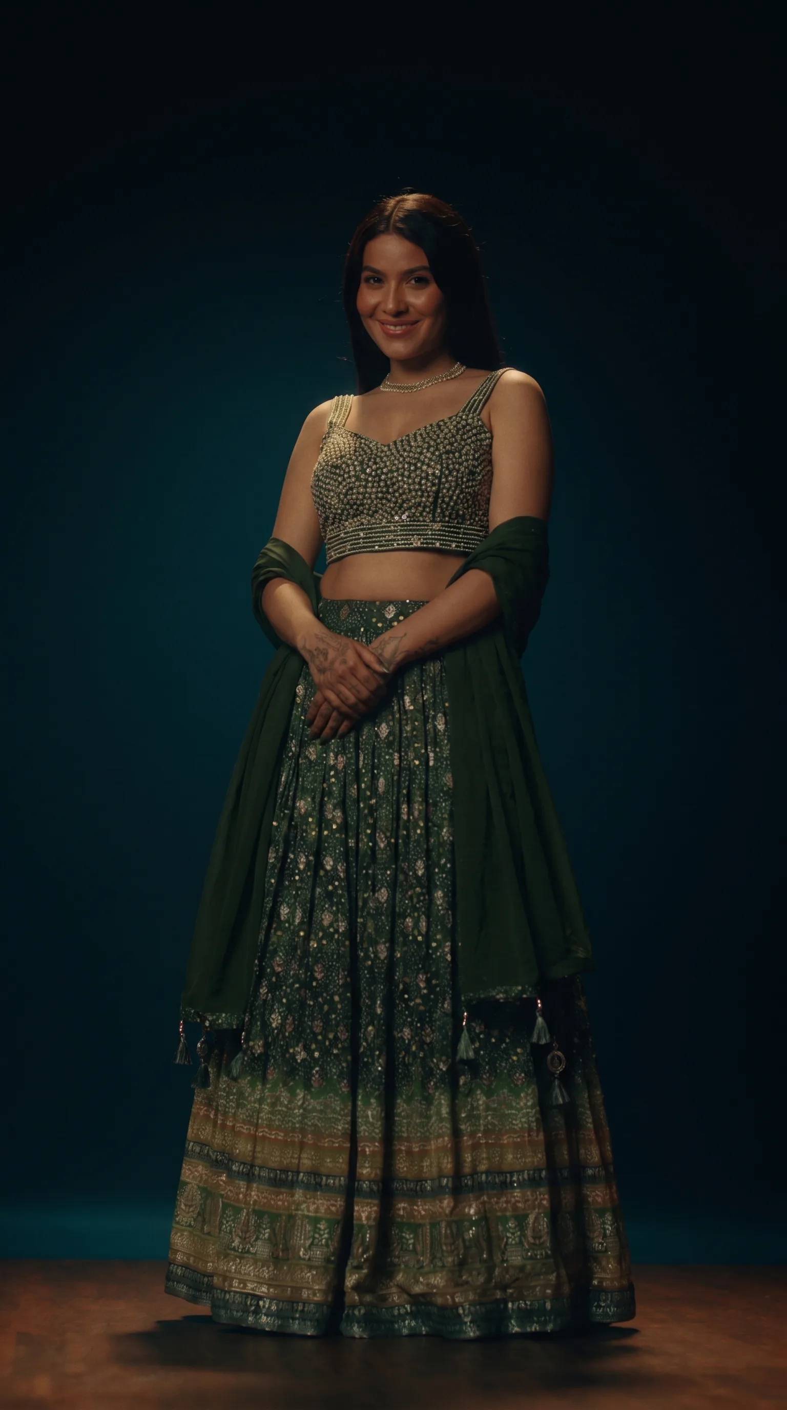 Front view of the green pearl-beaded ghaghara choli, woman standing with her hands clasped at the waist, warm key with the background falling to dark teal.