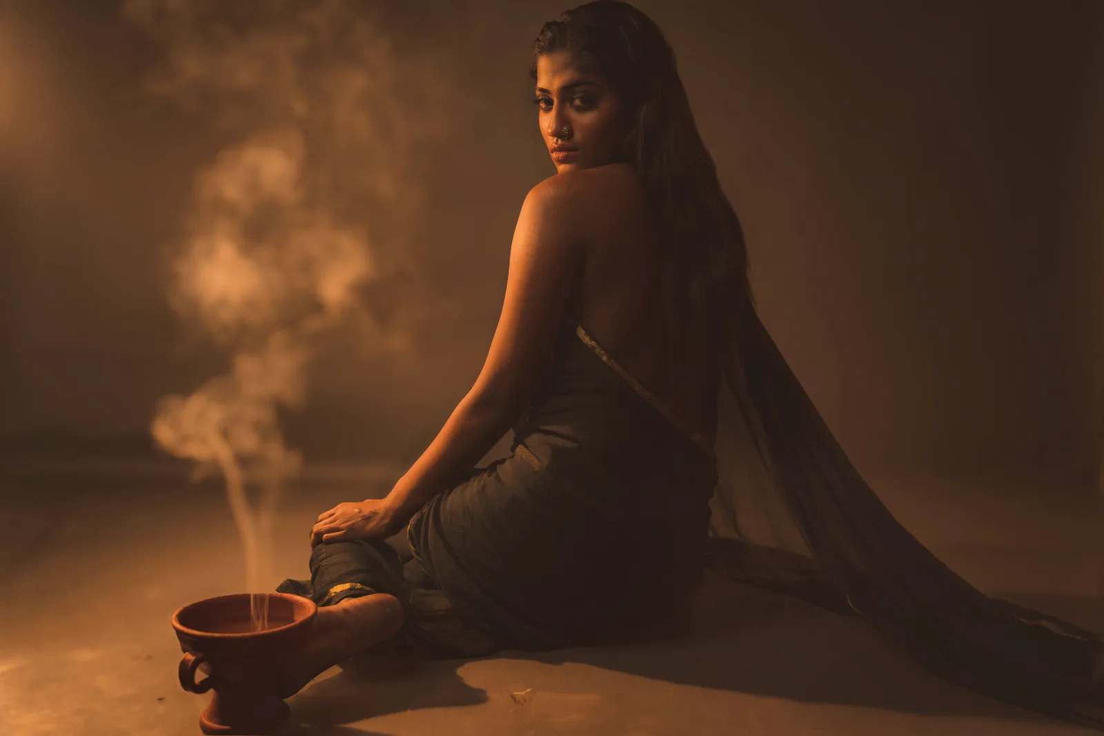 Close portrait of a woman holding the gold-trimmed edge of her black saree up over her shoulder, incense smoke rising beside her, looking at the camera.