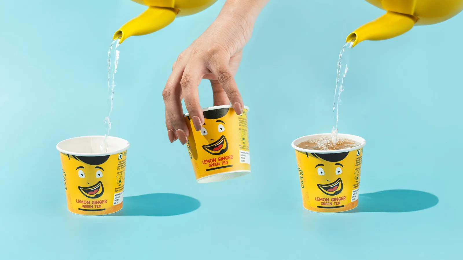 Yellow kettles pouring hot water into Lemon Ginger cups, one cup held mid-air by a hand, against a blue background.