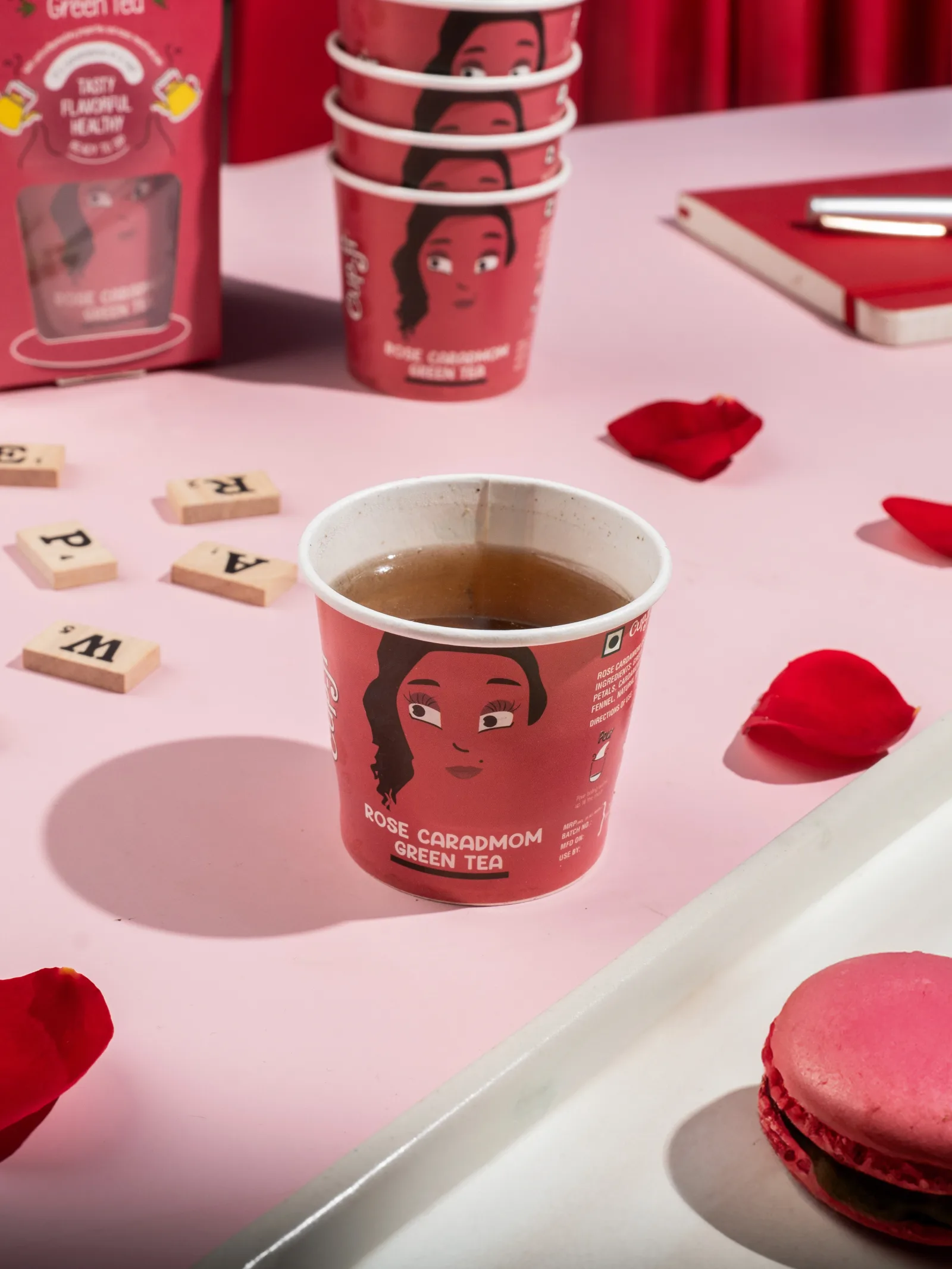Rose Cardamom cup on a pink desk with scrabble tiles, loose rose petals, a macaron and a stack of empty cups.