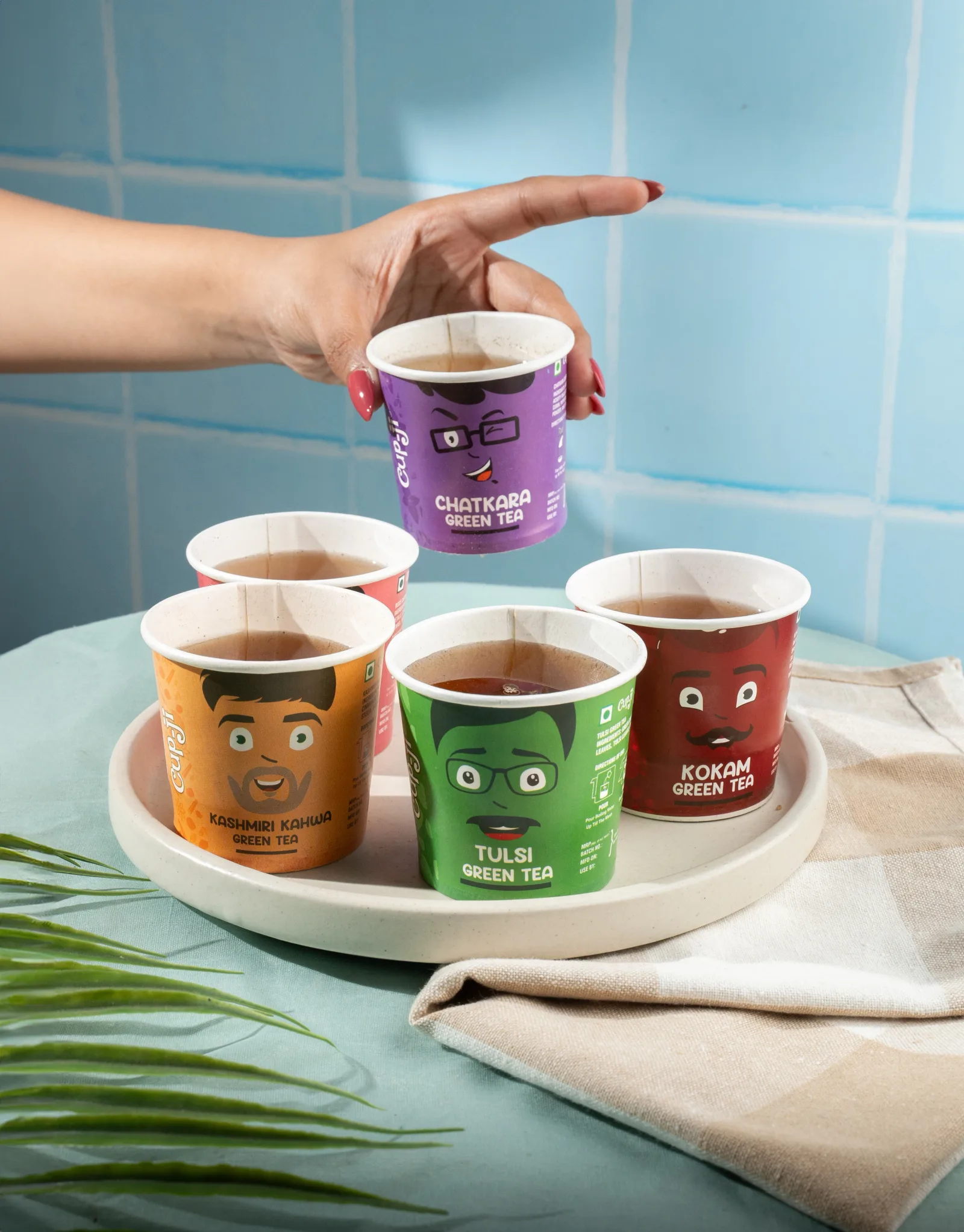 A hand lowering a Chatkara cup onto a tray already holding five filled cups, against a blue tiled wall with a palm leaf.