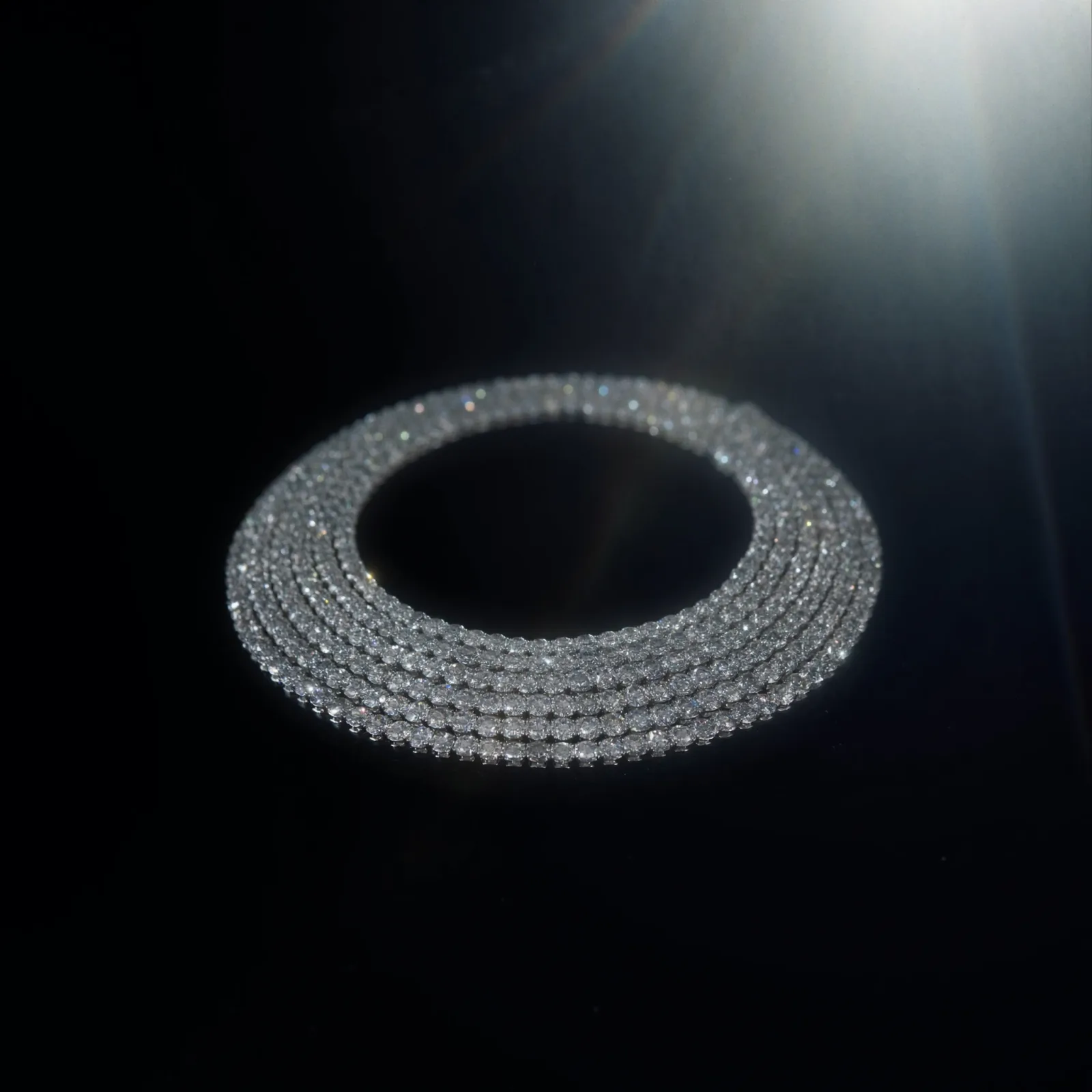 Diamond tennis necklace coiled on a dark glossy surface, rows of prong-set stones curving back on themselves.