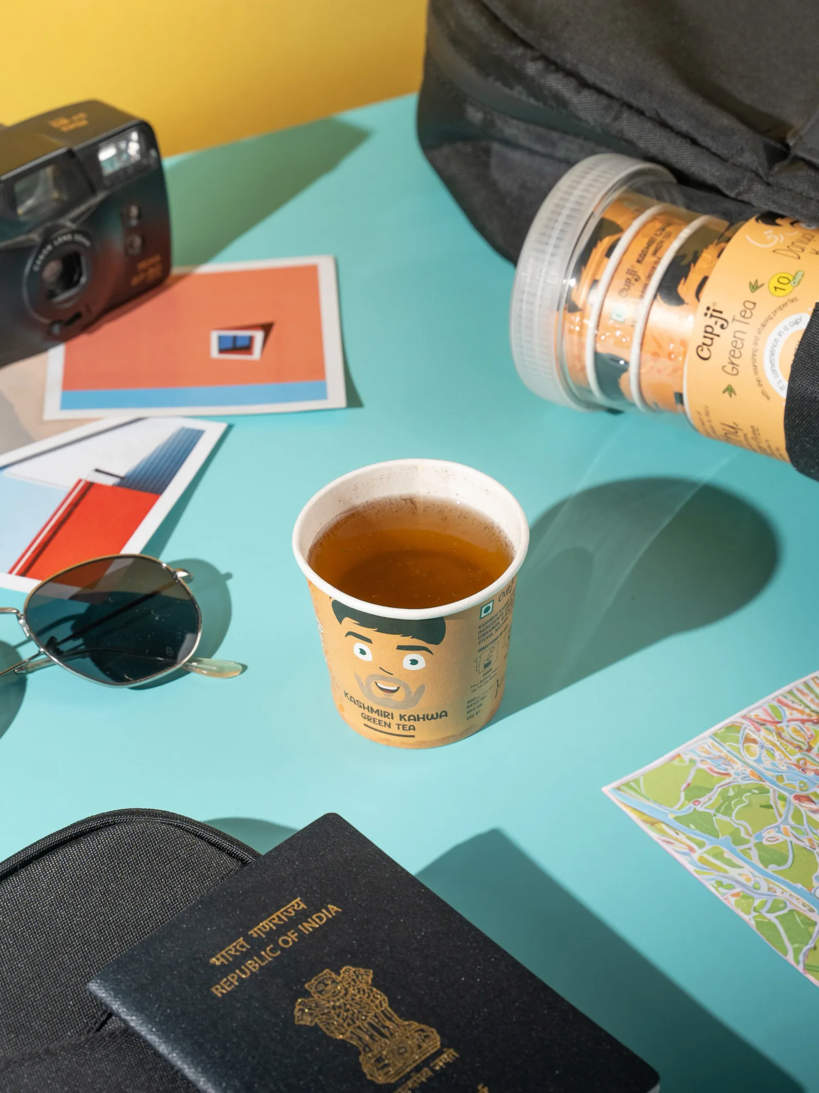 Travel flatlay with a passport, film camera, sunglasses and a Kashmiri Kahwa cup.
