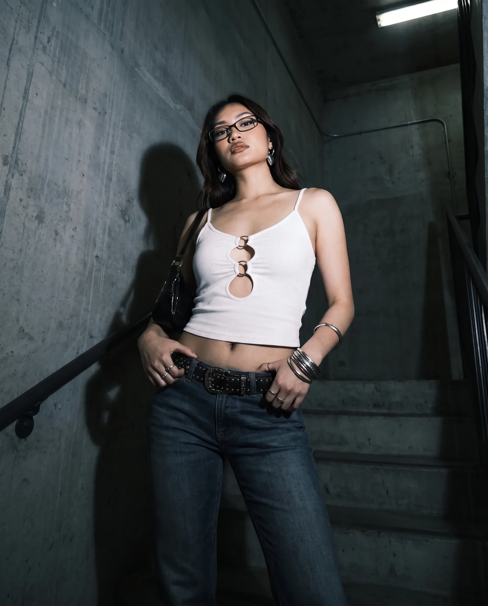 White ring-cutout cami, studded belt and jeans shot from a low angle in a concrete stairwell under a strip light.