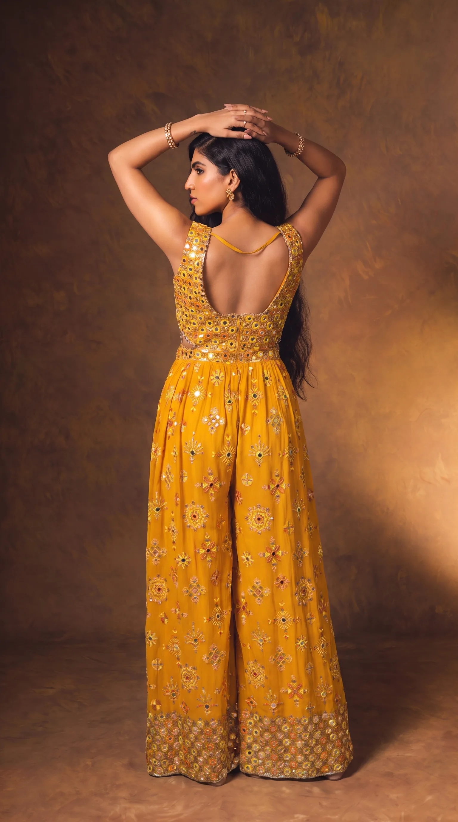 Woman in a mustard mirror-work sleeveless jumpsuit with wide palazzo legs, both arms raised overhead so the open back is visible, deep shadow holding to the left of frame.
