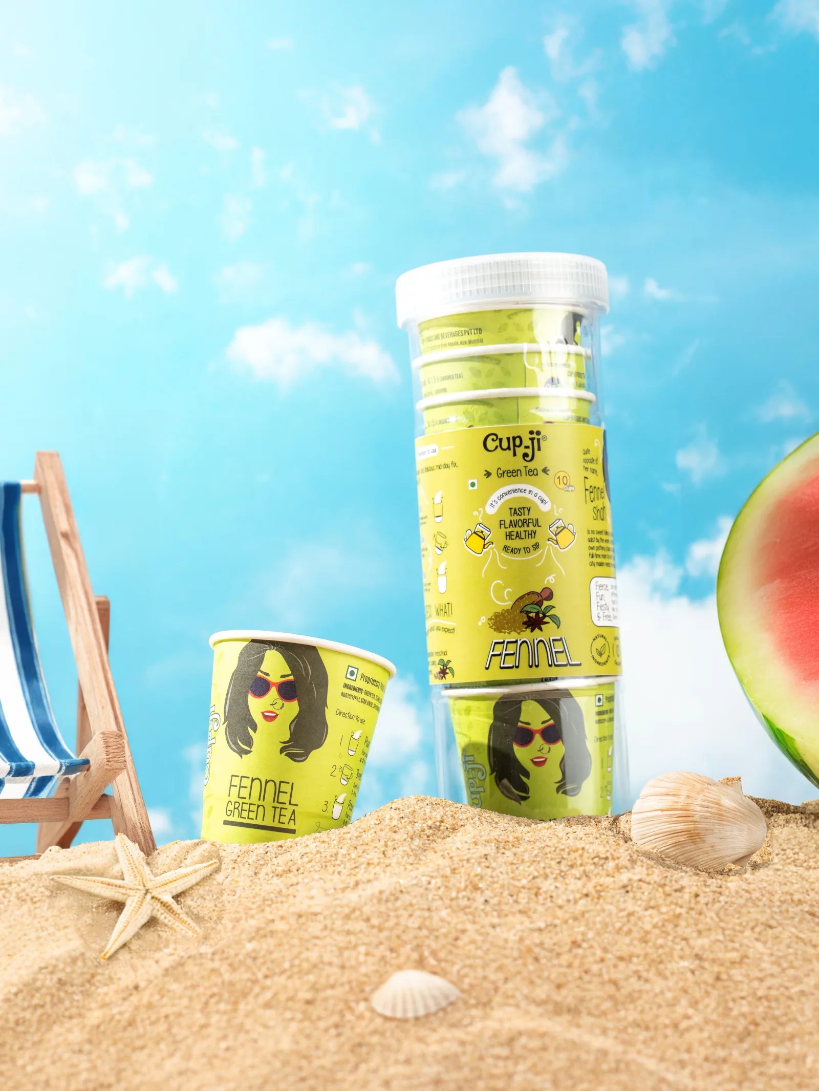 Summer campaign shot: Fennel jar and cup staged as a beach scene with a miniature deck chair, watermelon, shells and sand.