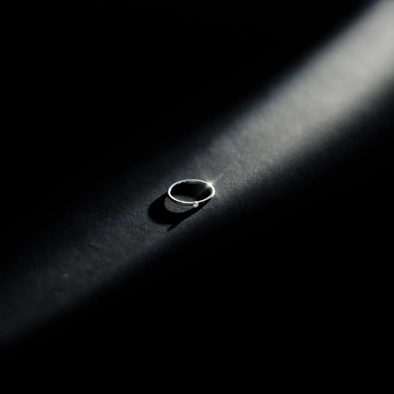 Slim diamond solitaire ring standing upright against black under a narrow shaft of light.