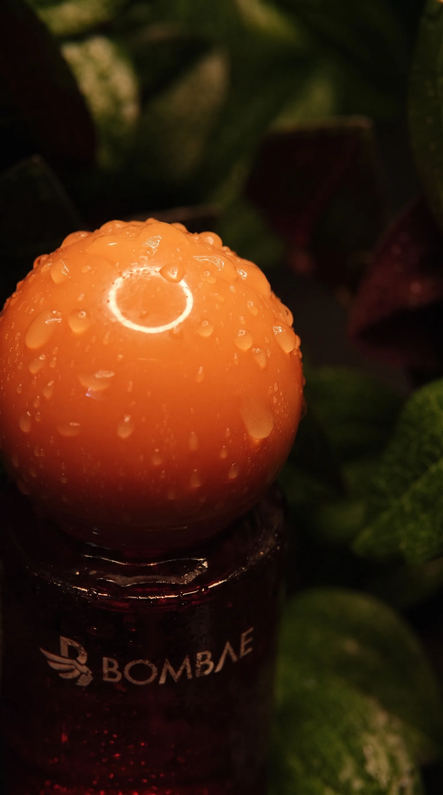 Top-down macro of the Bombae Maya bottle's orange cap covered in water droplets with a bright specular highlight ring.