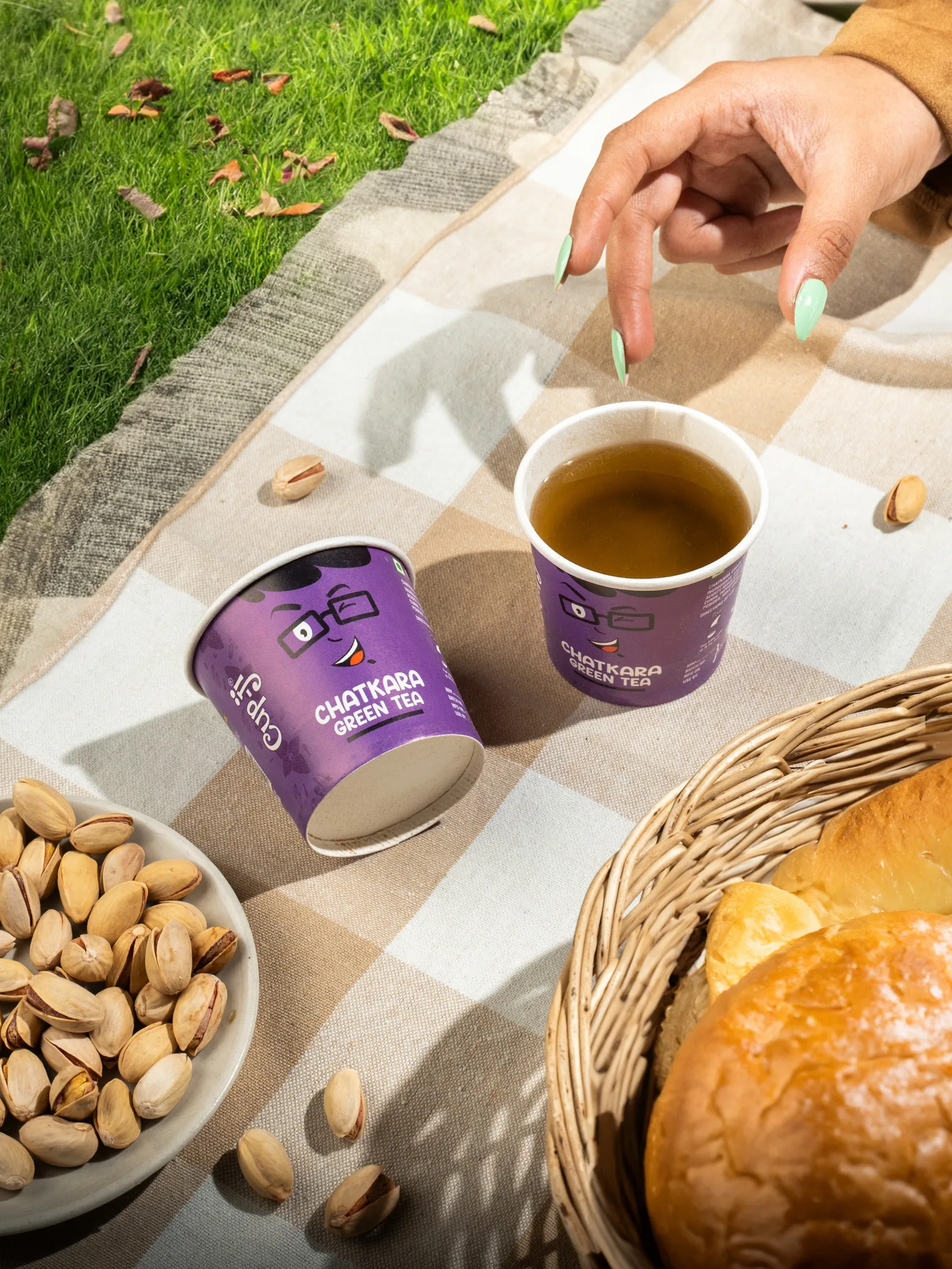 Two Chatkara cups on a picnic blanket on grass with pistachios and a bread basket, a hand reaching for one.