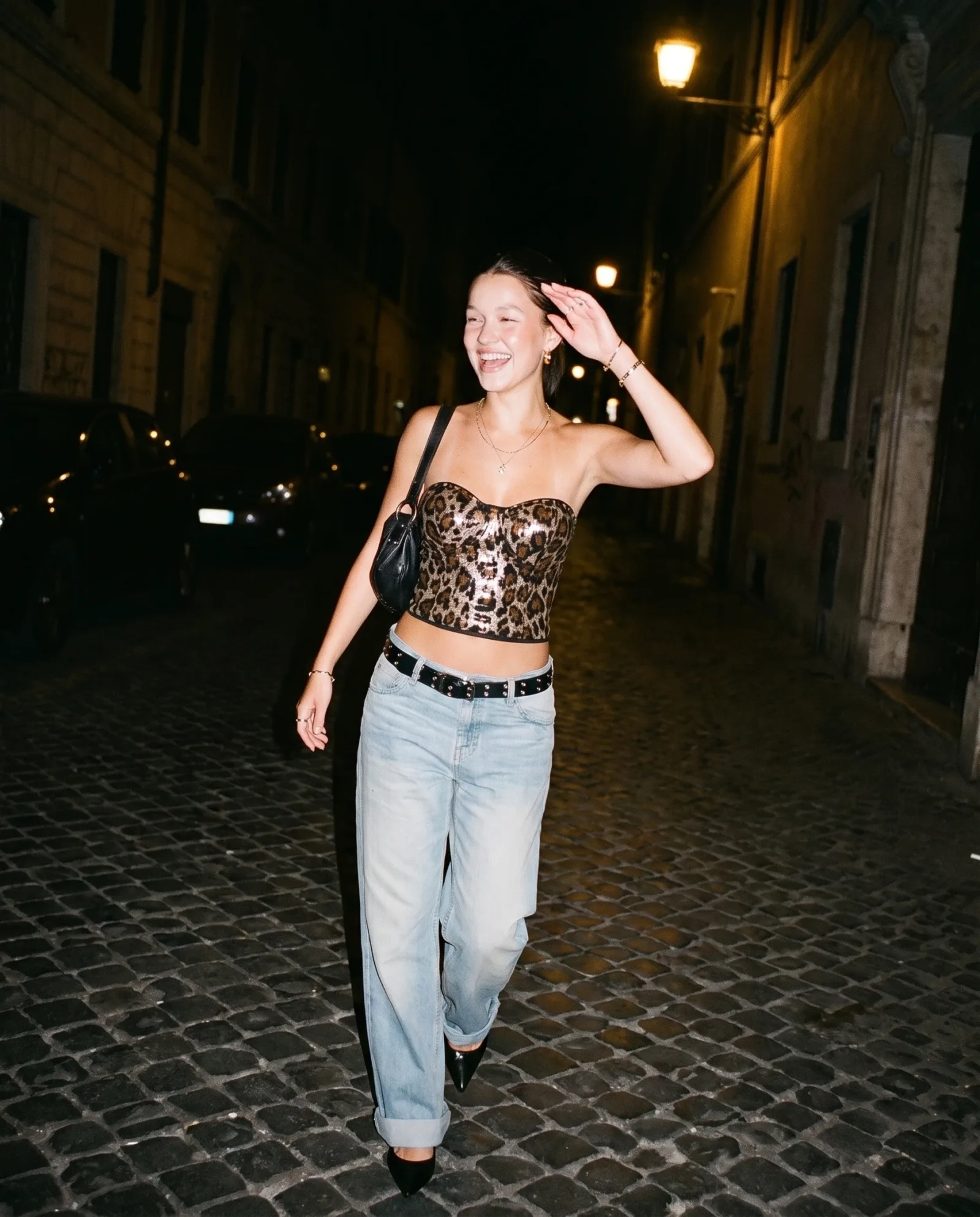 Leopard sequin bustier with low-rise jeans and a studded belt, walking a cobbled street at night, hand lifted to the hair.