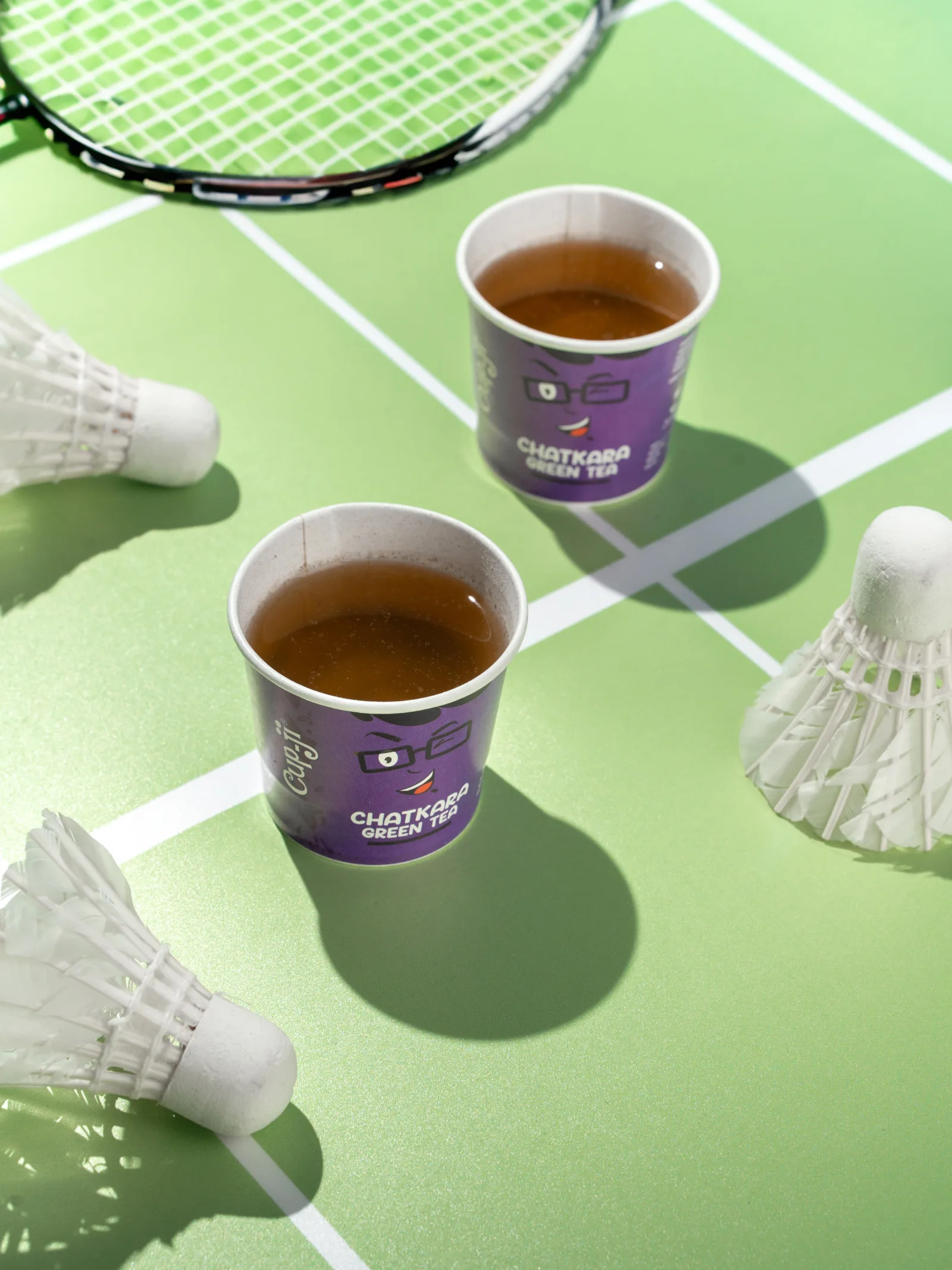 Two Chatkara cups on a green badminton court beside a racket and shuttlecocks.