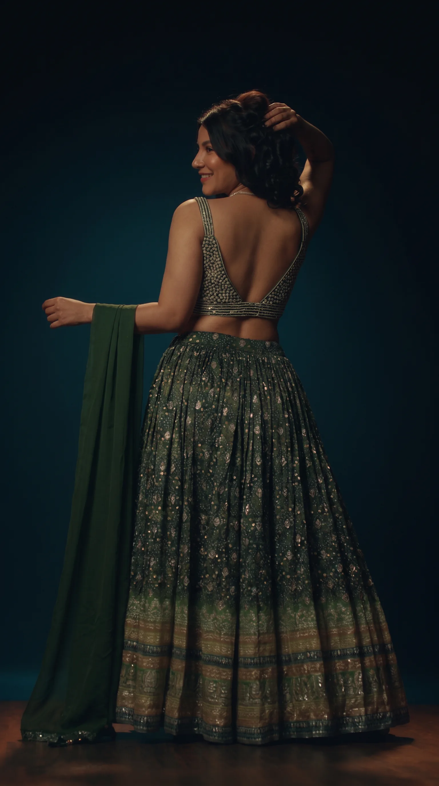 Woman in a green pearl-beaded ghaghara choli seen from behind, one arm raised to her hair and the other holding the dupatta, looking back over her shoulder against a dark teal background.