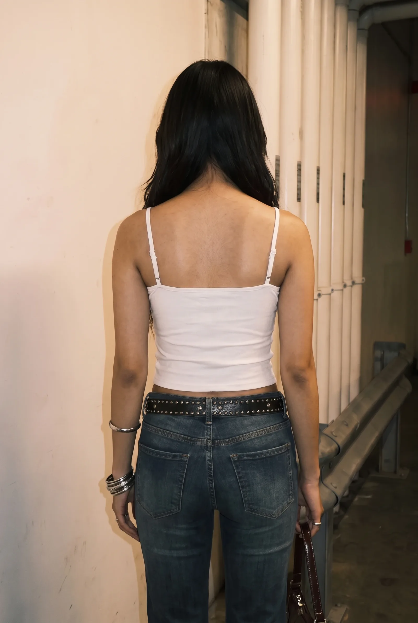 Back view of the white ring-cutout cami with a studded belt and jeans, walking down a service corridor lined with pipes.