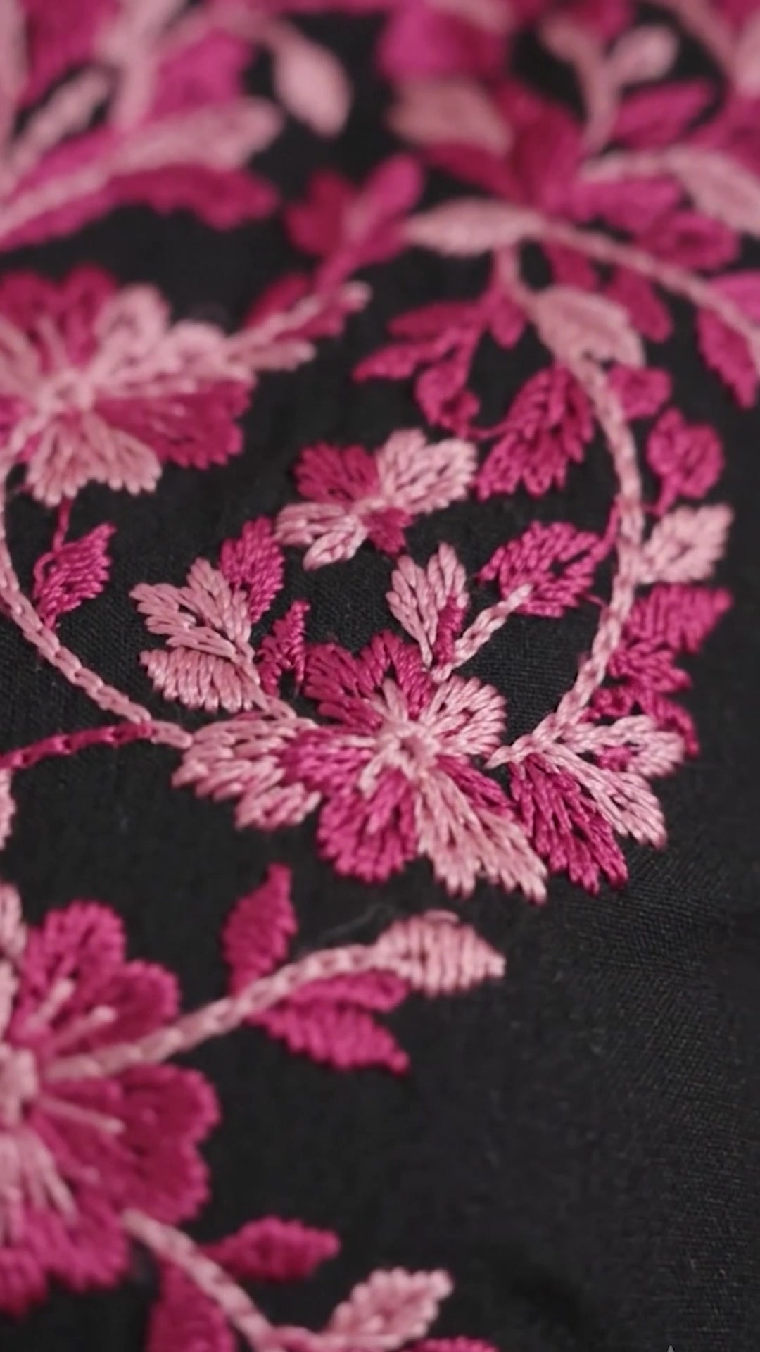 Full-length vertical film of a black kurti with pink floral chikankari across the yoke and sleeves, worn with dark denim and silver jhumka earrings against a beige wall.