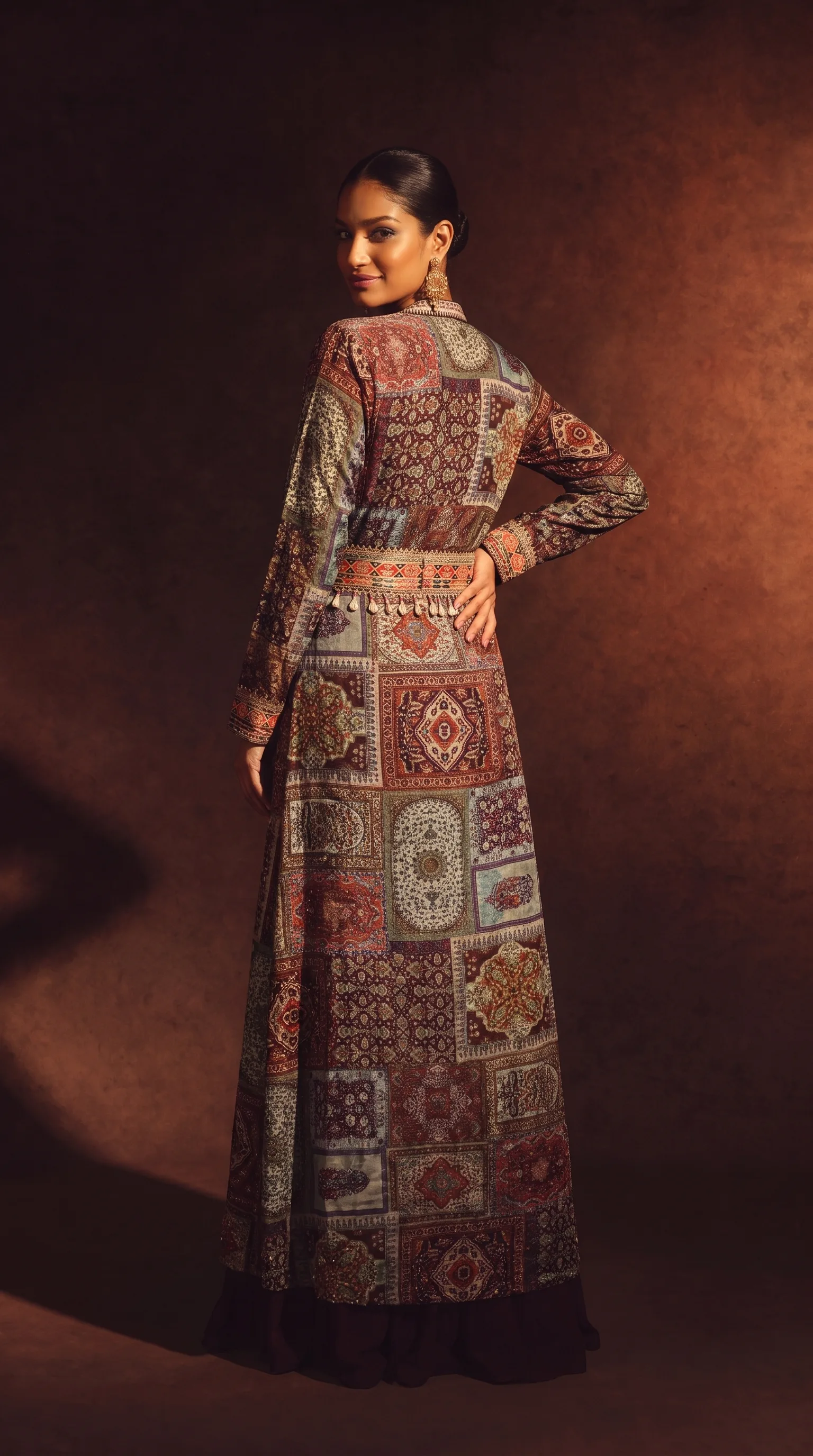 Back view of the same patchwork shrug gown, the woman glancing over her shoulder, the full embroidered back panel visible, deep shadow holding to the left of frame.