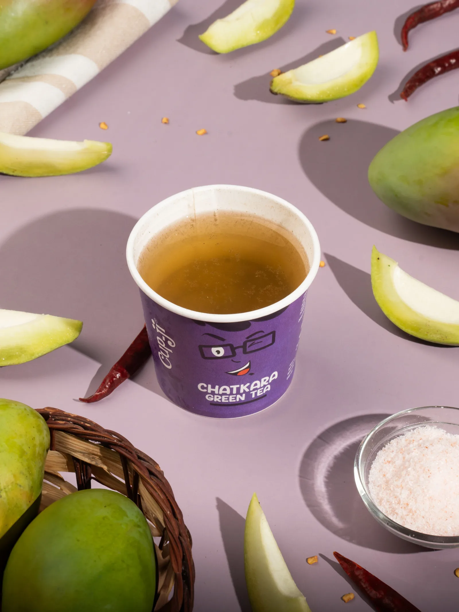 Chatkara cup with raw mango wedges, red chilli and pink rock salt on a lavender surface.
