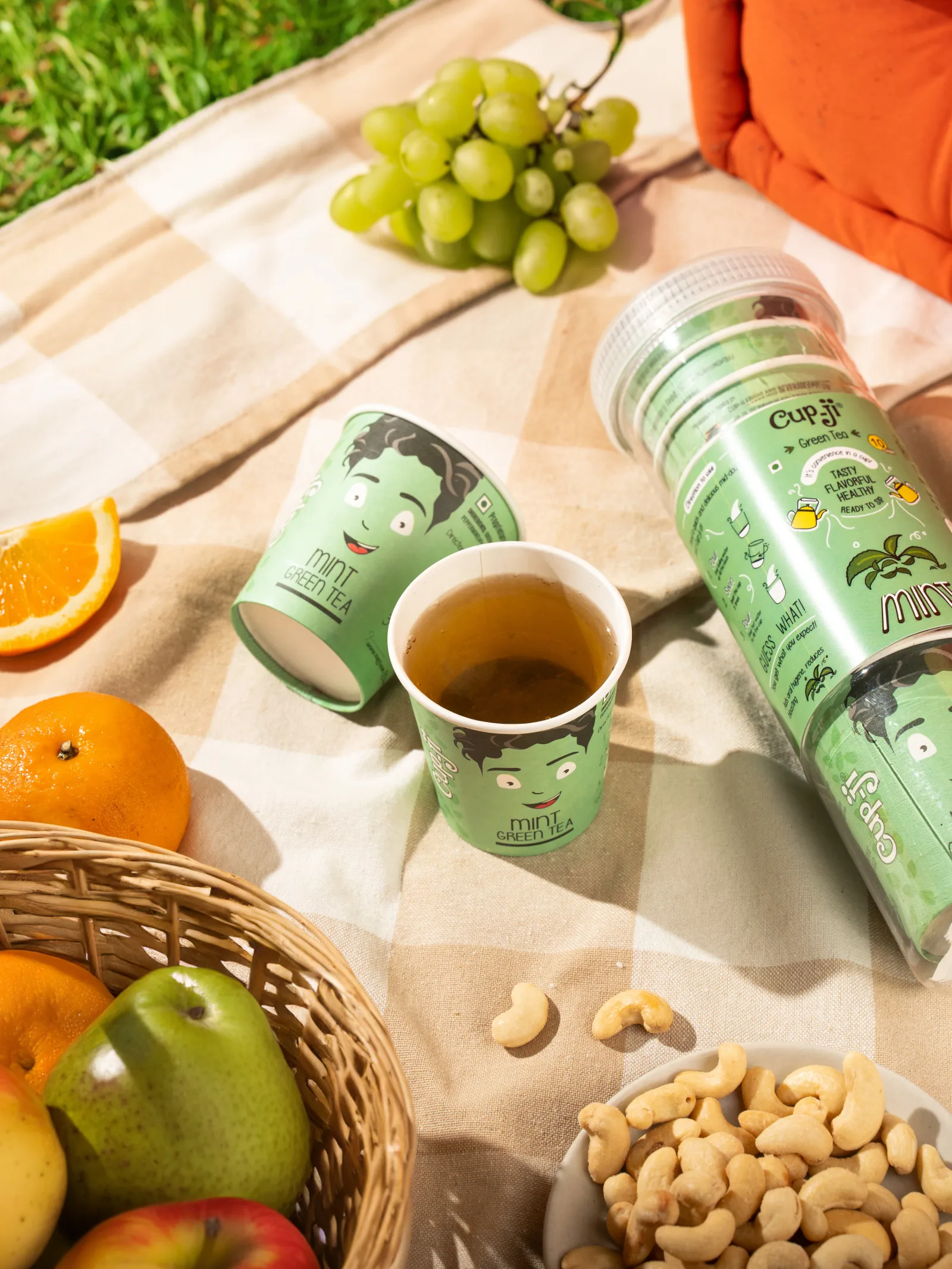 Summer campaign shot: Mint Green Tea cup on a picnic blanket with grapes, citrus and cashews.