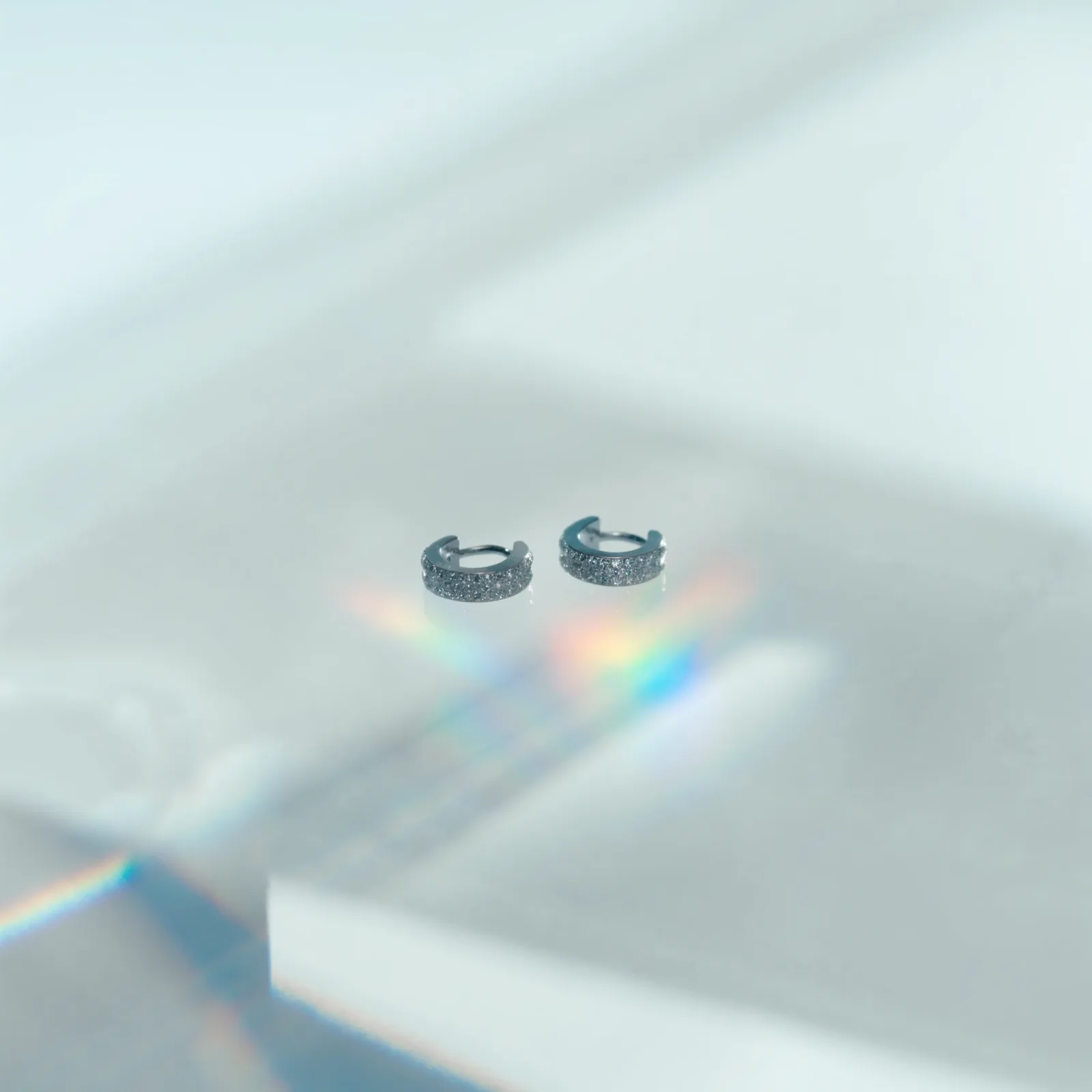 Pair of pavé diamond huggie hoop earrings on a white surface with a spectrum cast beside them.