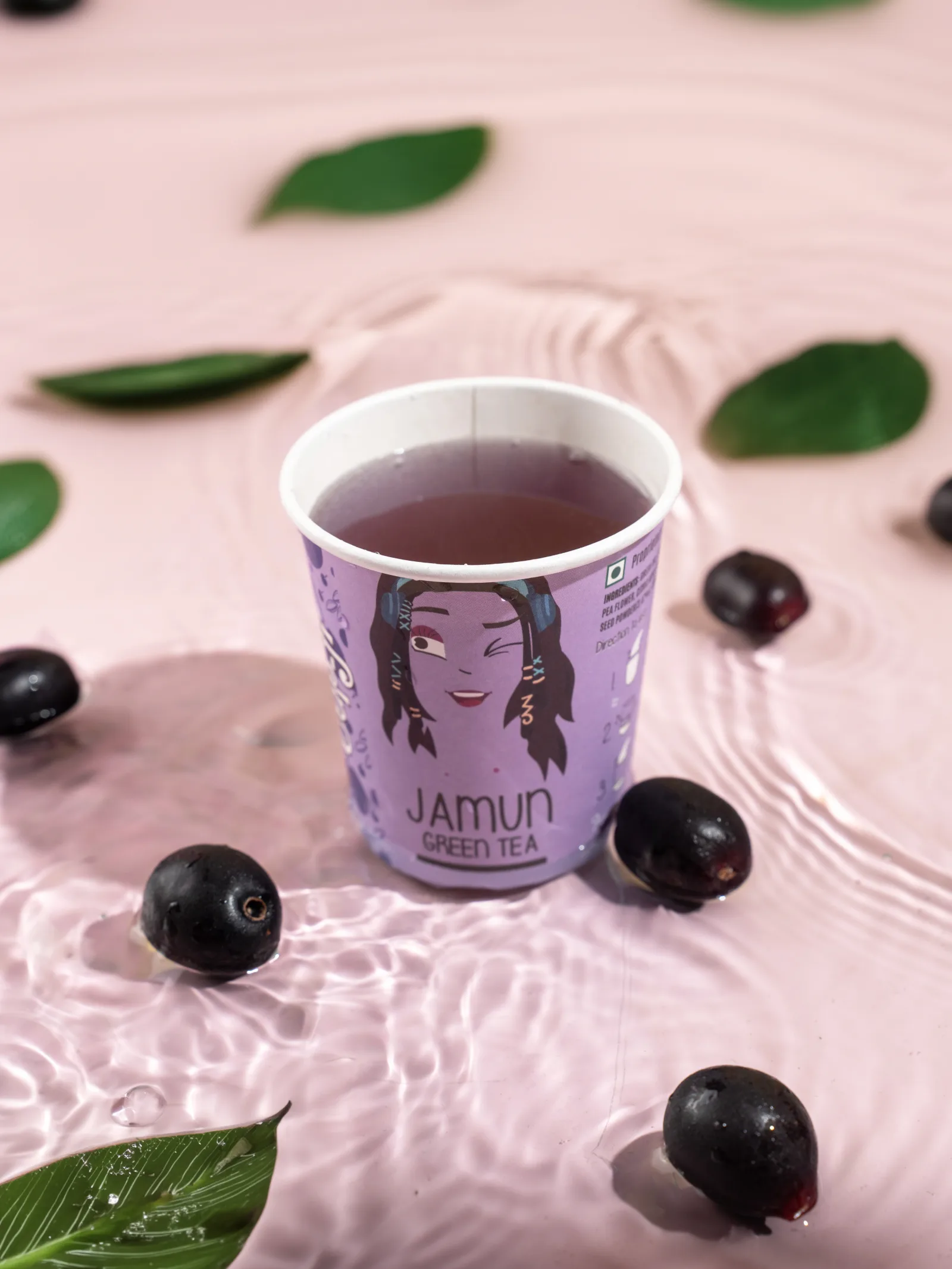 Jamun tea being poured into a cup on a wet pink surface, whole jamun fruit and leaves in close focus.
