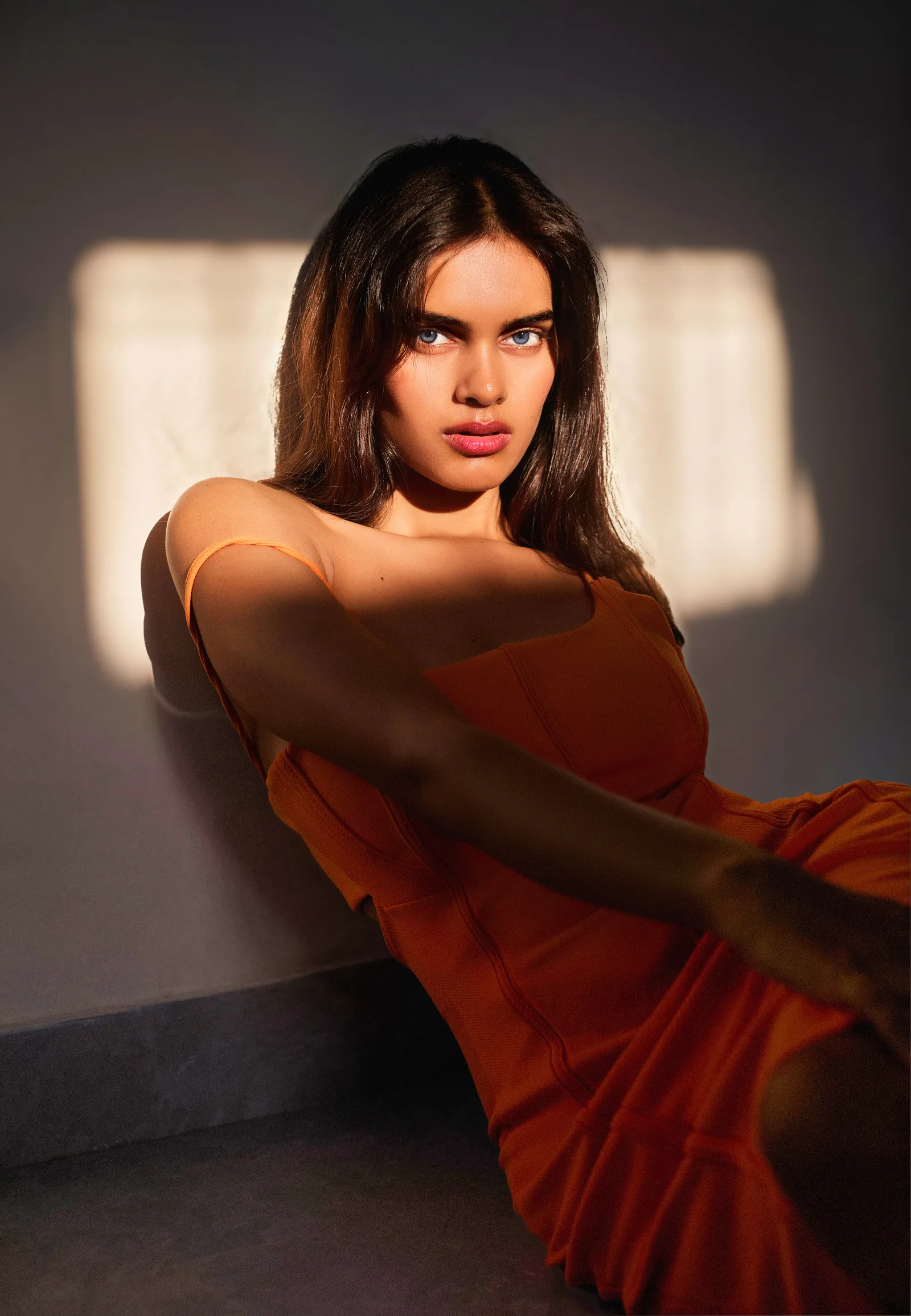 Woman in an orange dress seated against a wall, hard window light casting blind-slat shadows beside her, looking at the camera with one arm raised.