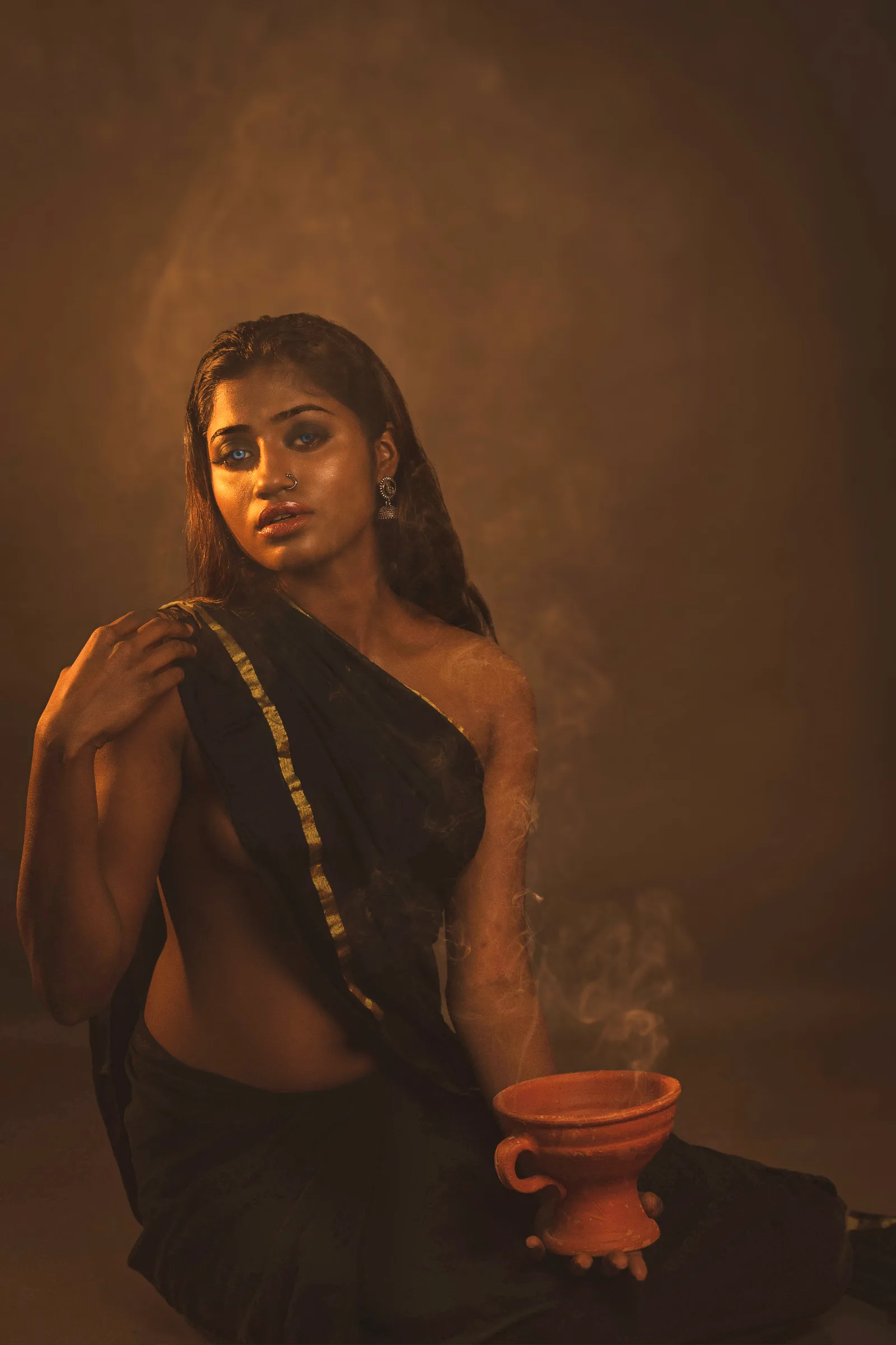 Woman photographed from the side and back, holding a smoking clay pot close to her face, saree slipping off one shoulder, looking back at the camera.