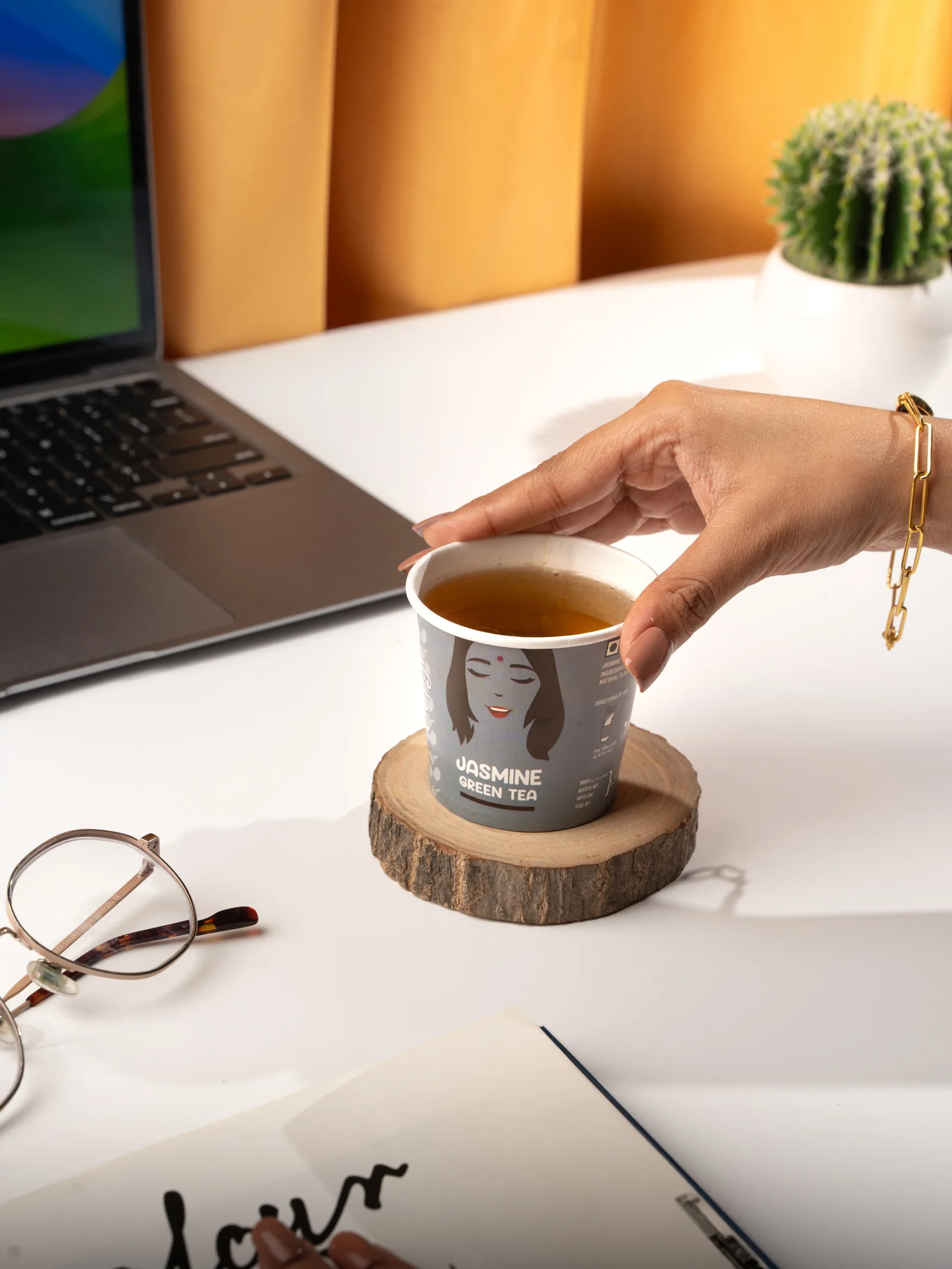 A hand lifting a Jasmine cup off a wooden coaster on a working desk with a laptop, glasses and a small cactus.
