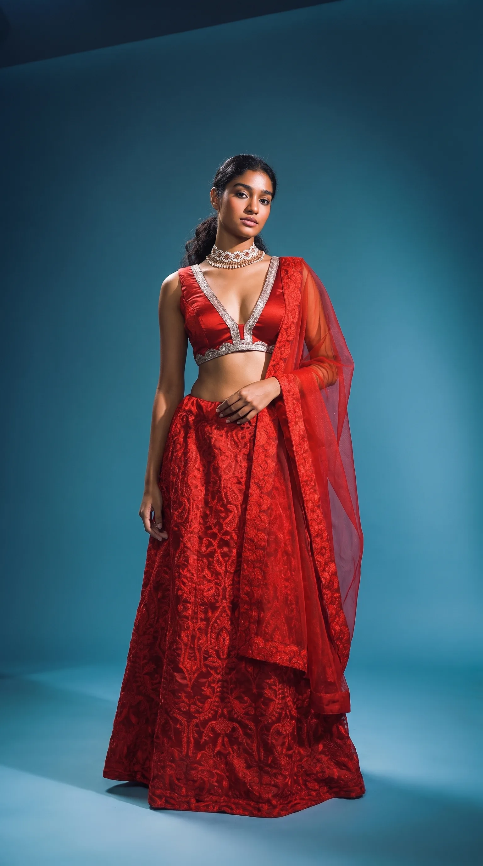 Woman in a red silk-blend thread-work lehenga, body turned with the net dupatta caught mid-sweep, warm key from camera-right against a teal gradient.