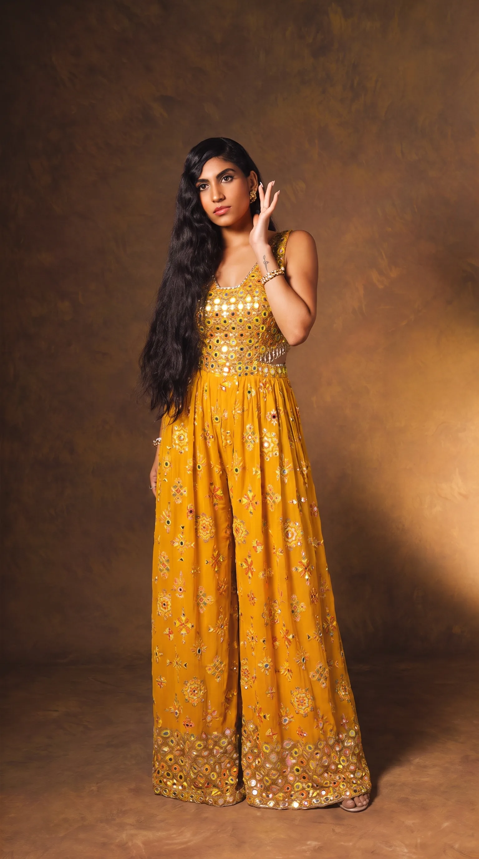 Front view of the mustard mirror-work jumpsuit, woman with her chin up and one hand to her temple, allover mirror embroidery catching a warm directional light.