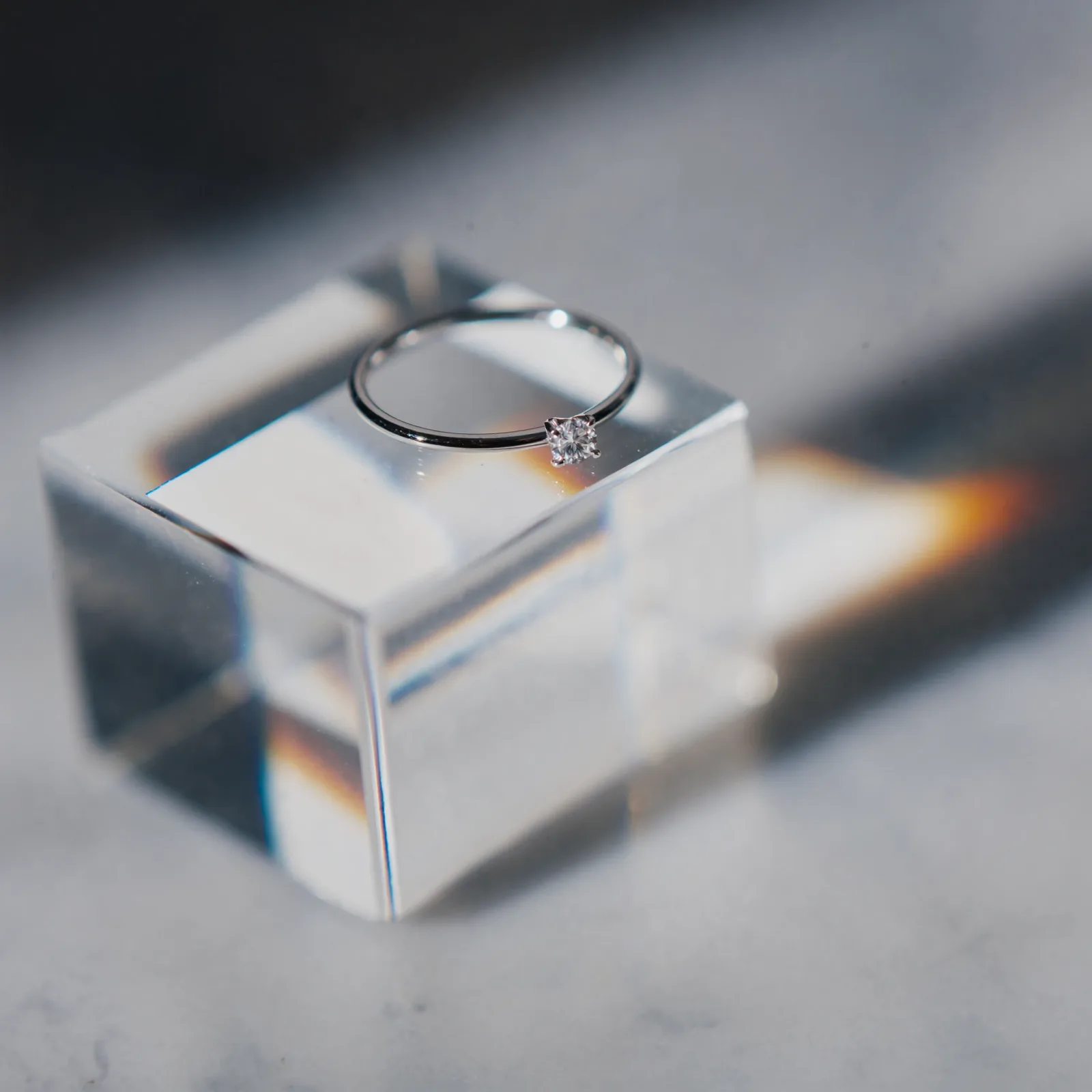 Round brilliant diamond solitaire ring standing on a clear crystal block, a prism casting a rainbow across the surface.