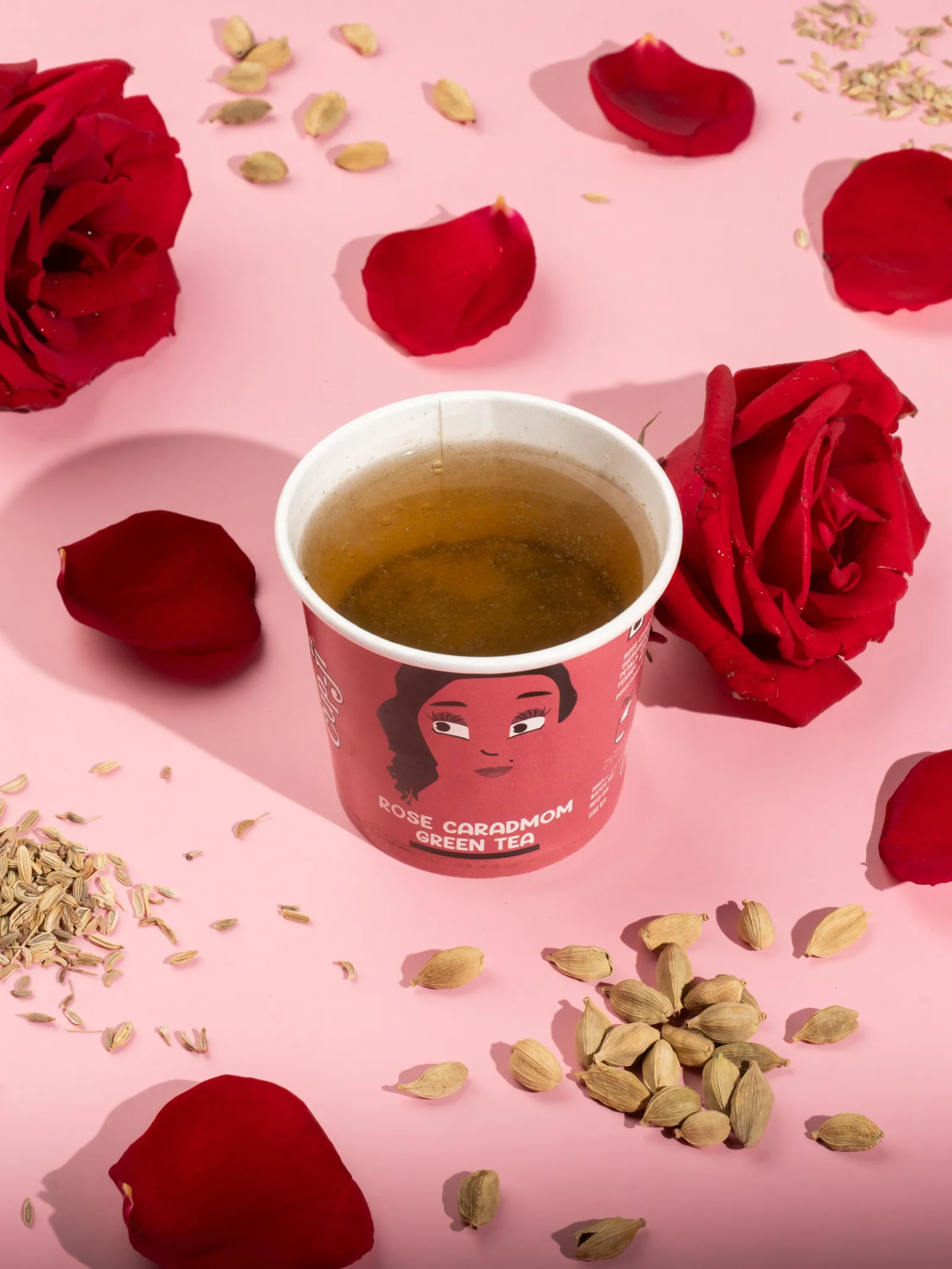 Rose Cardamom cup with whole roses, loose petals and green cardamom pods on a pink surface.