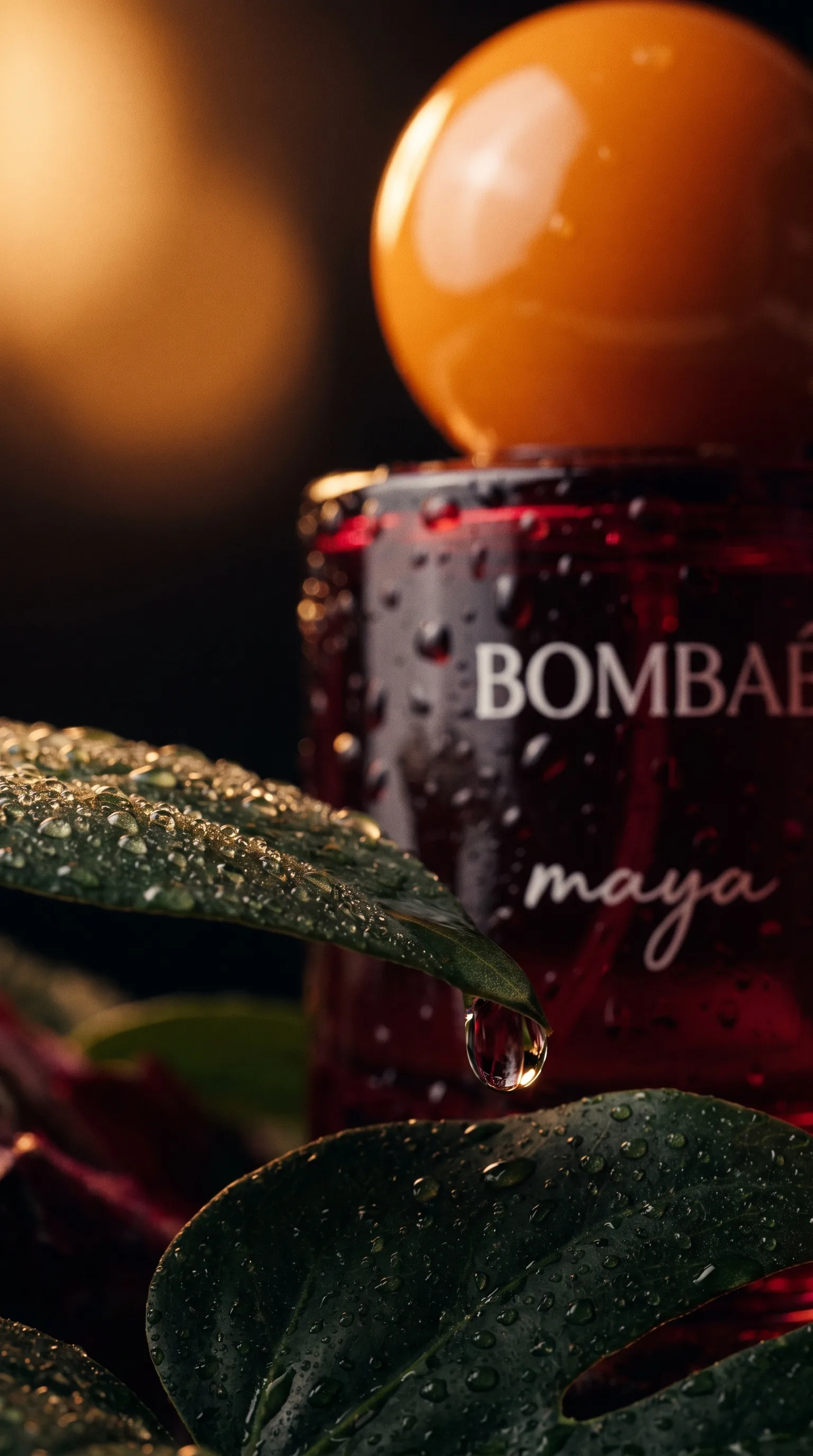 Water droplet hanging from a wet leaf tip in front of the blurred Bombae Maya bottle, its orange cap glowing out of focus behind.