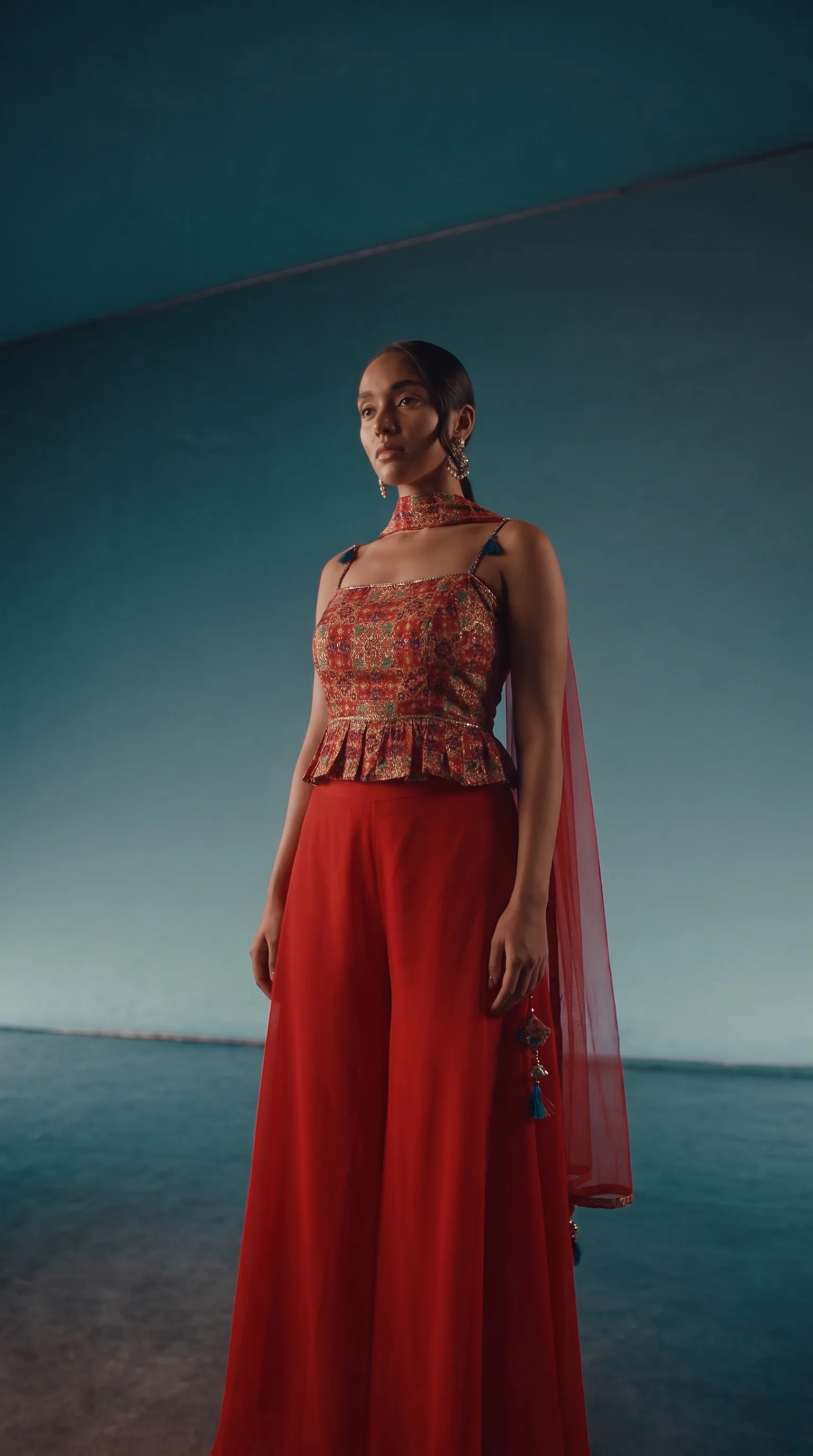 Woman in a multicolour patchwork peplum top with tasselled halter straps and wide red palazzo, one hand at her shoulder looking away, standing in an architectural tunnel under a teal-to-dark gradient sky.