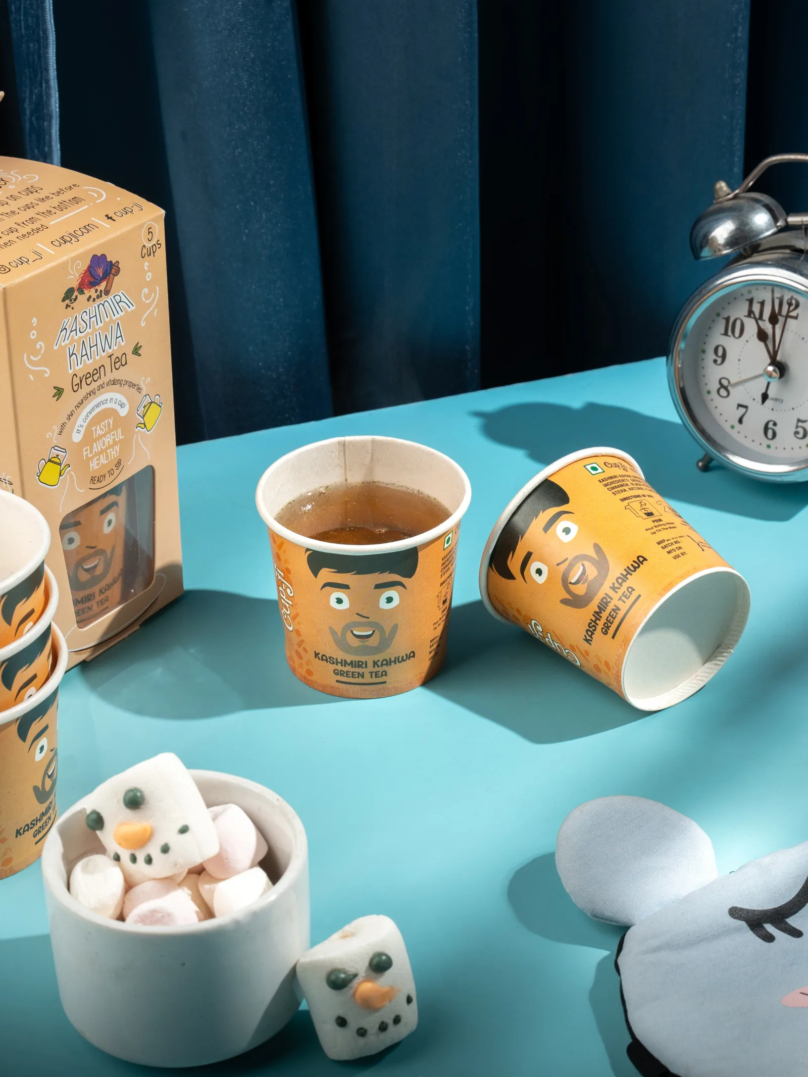 Kashmiri Kahwa cups on a teal table, one tipped on its side, with an alarm clock, a pack and marshmallow snowmen.