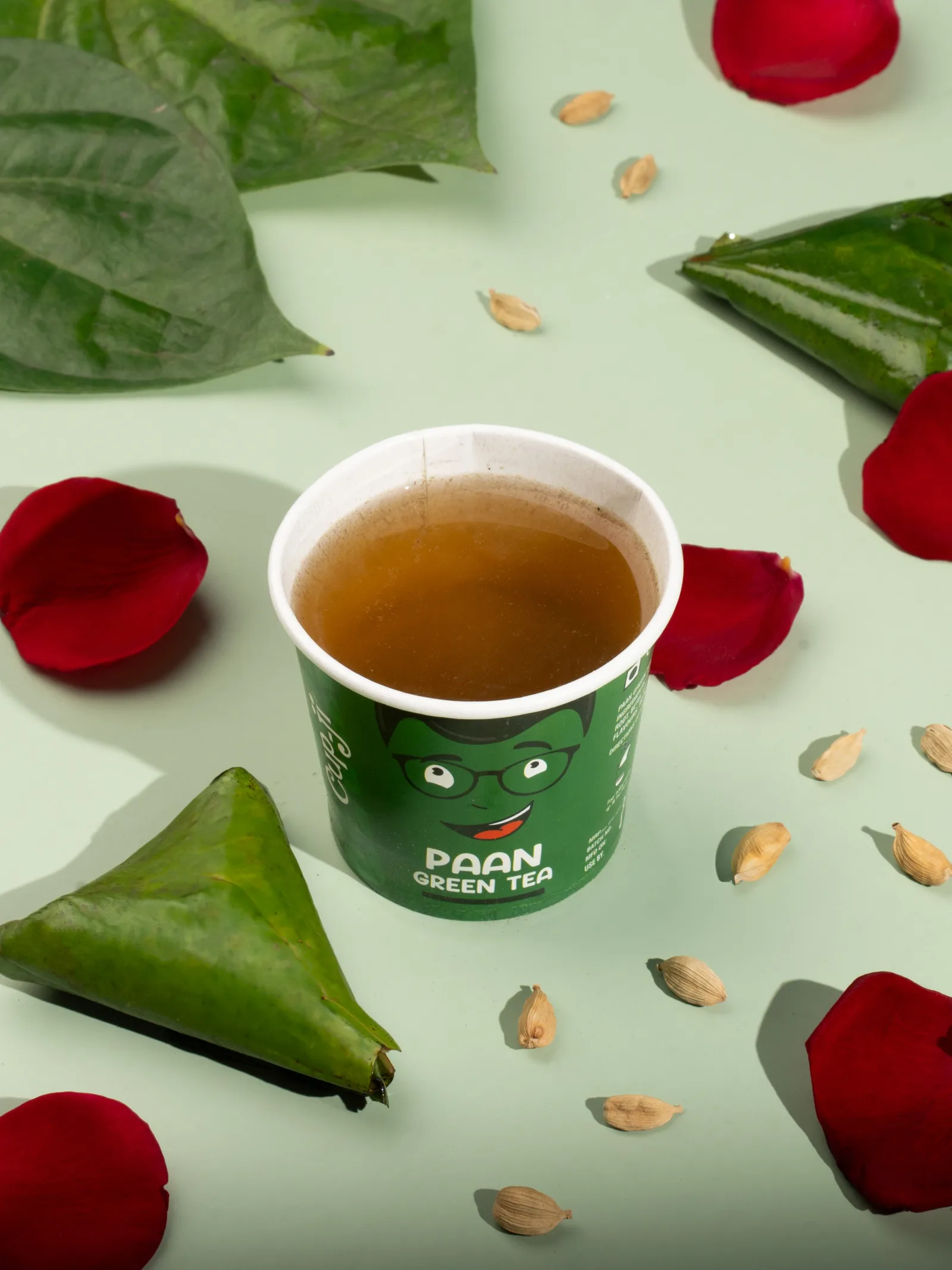 Paan cup with folded betel leaf parcels, cardamom pods and a rose petal on a mint green surface.