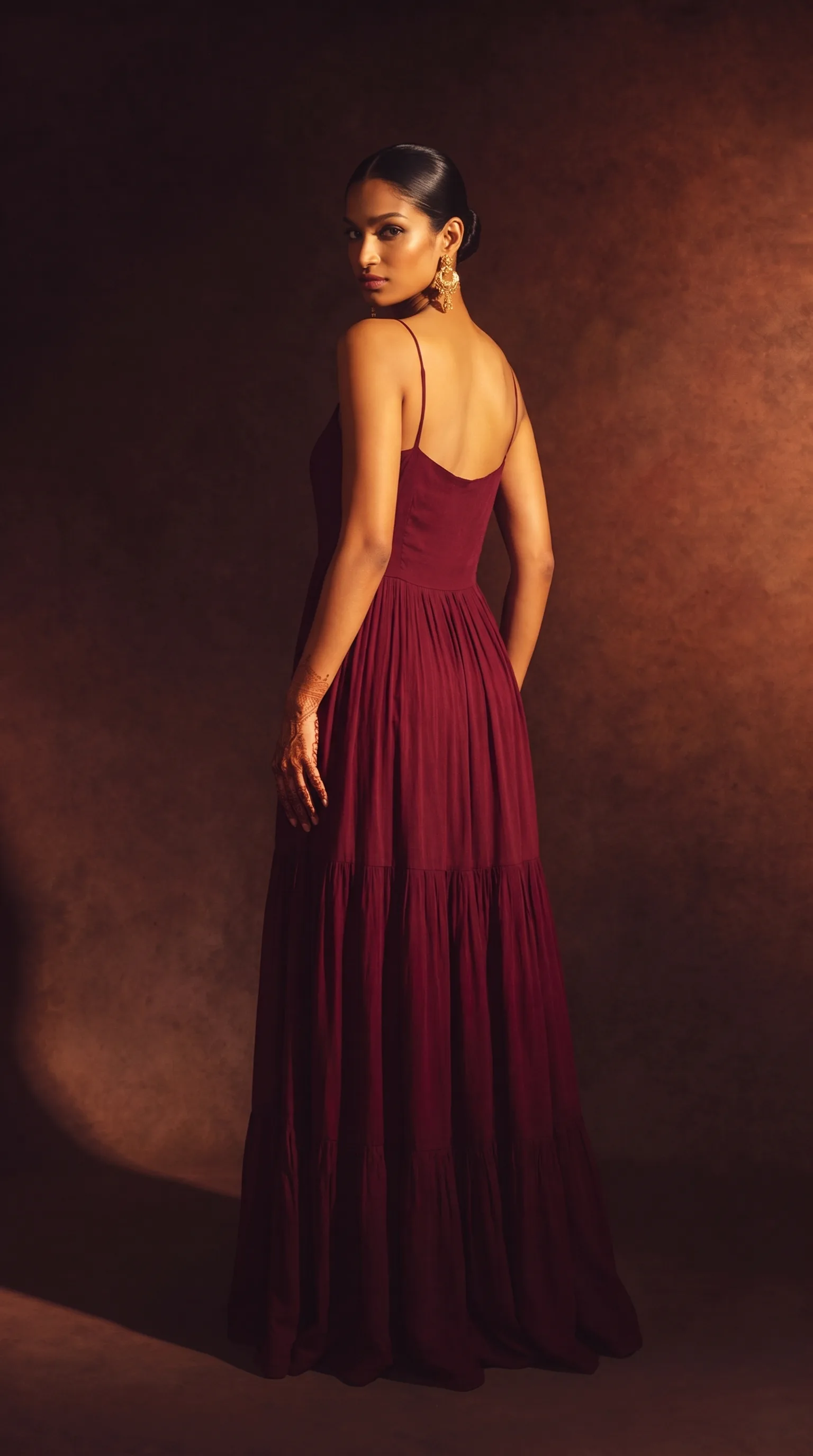 Woman in a full-length indo-western gown with a long patchwork shrug and a detachable tasselled belt, hands framing her face, lit from camera-right against a warm amber gradient backdrop.
