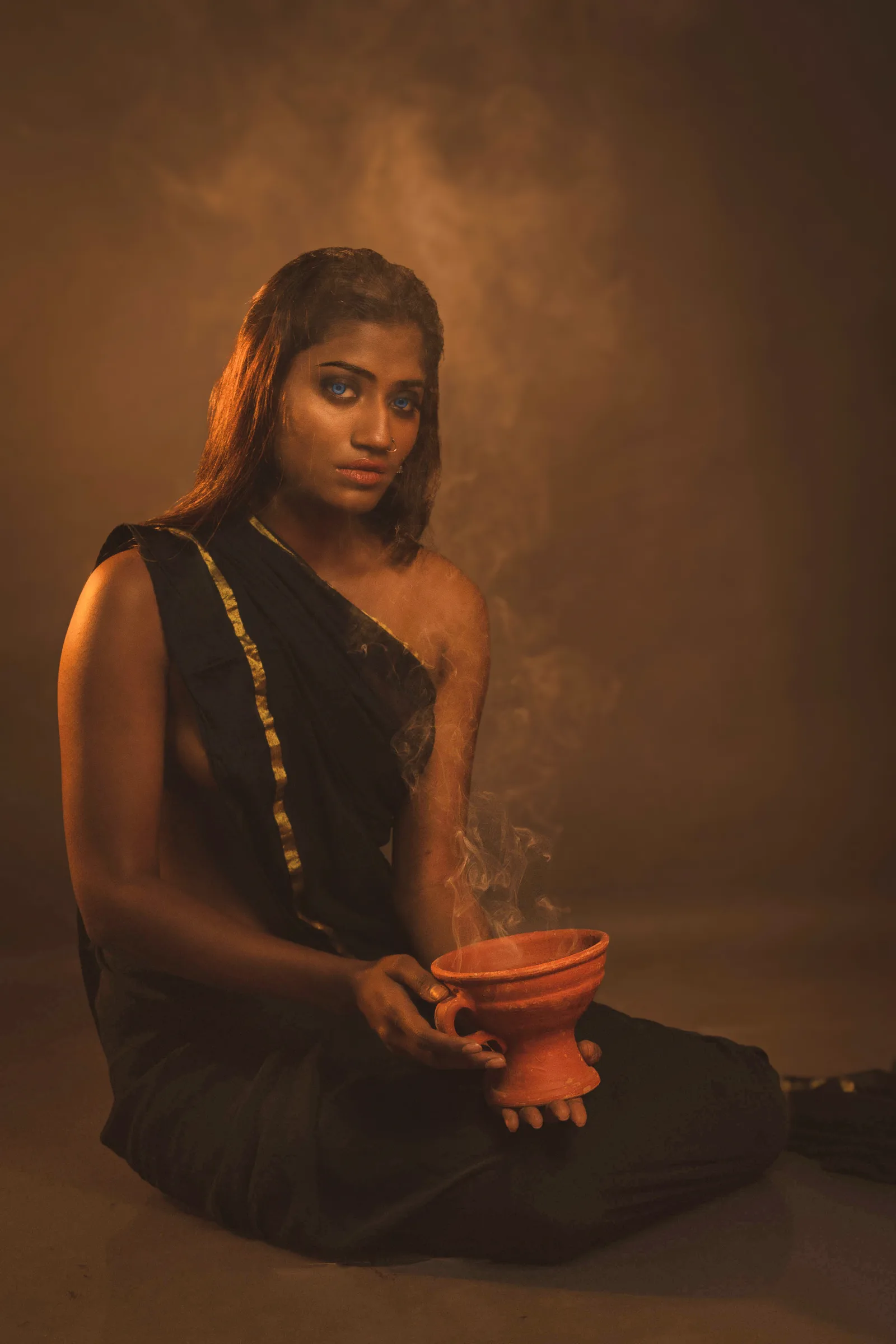 Woman seated with her back and bare shoulder to the camera, looking over her shoulder, a smoking clay pot on the floor beside her, sheer black drape trailing behind.