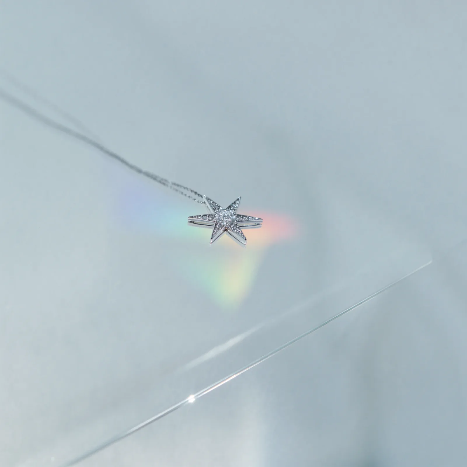 Pavé diamond star pendant on a fine chain, lying on a pale reflective surface with a soft spectrum beside it.