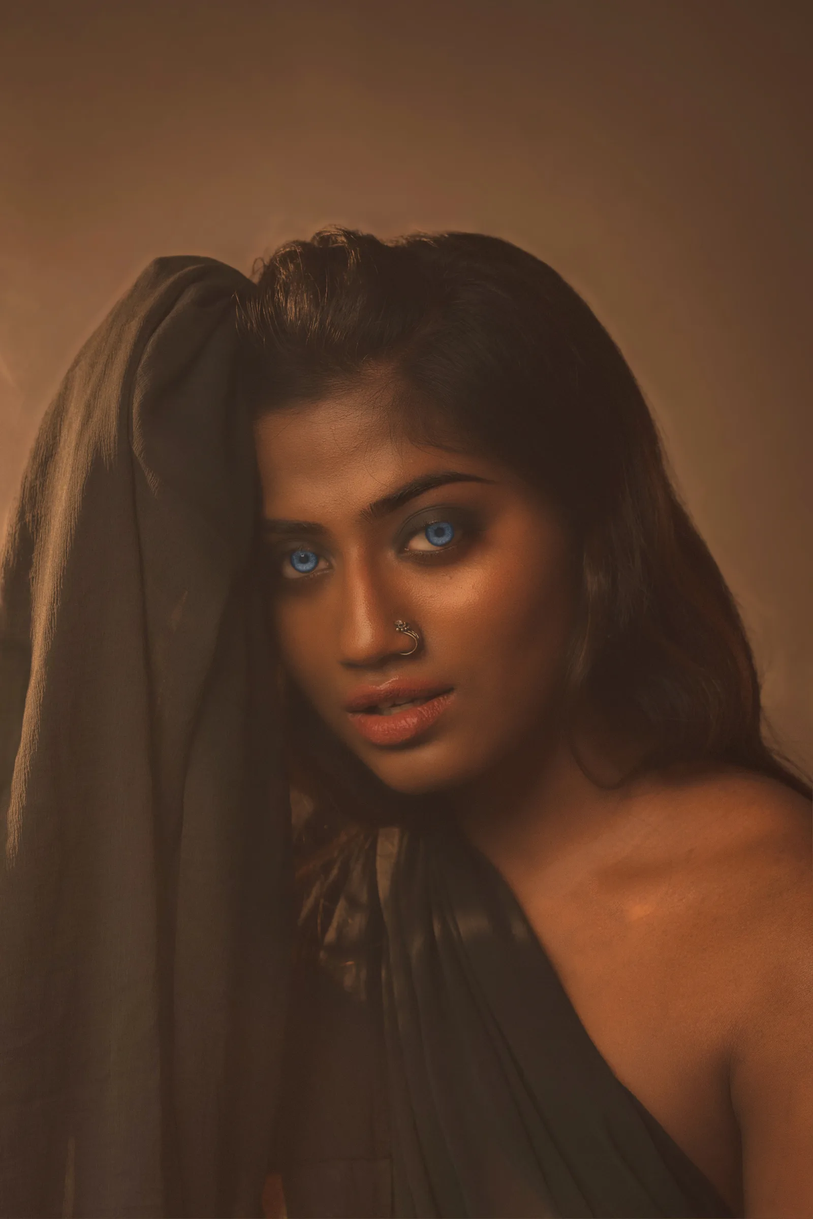 Woman seated cross-legged in a gold-trimmed black saree, holding a smoking clay incense pot, looking directly at the camera in warm amber light.