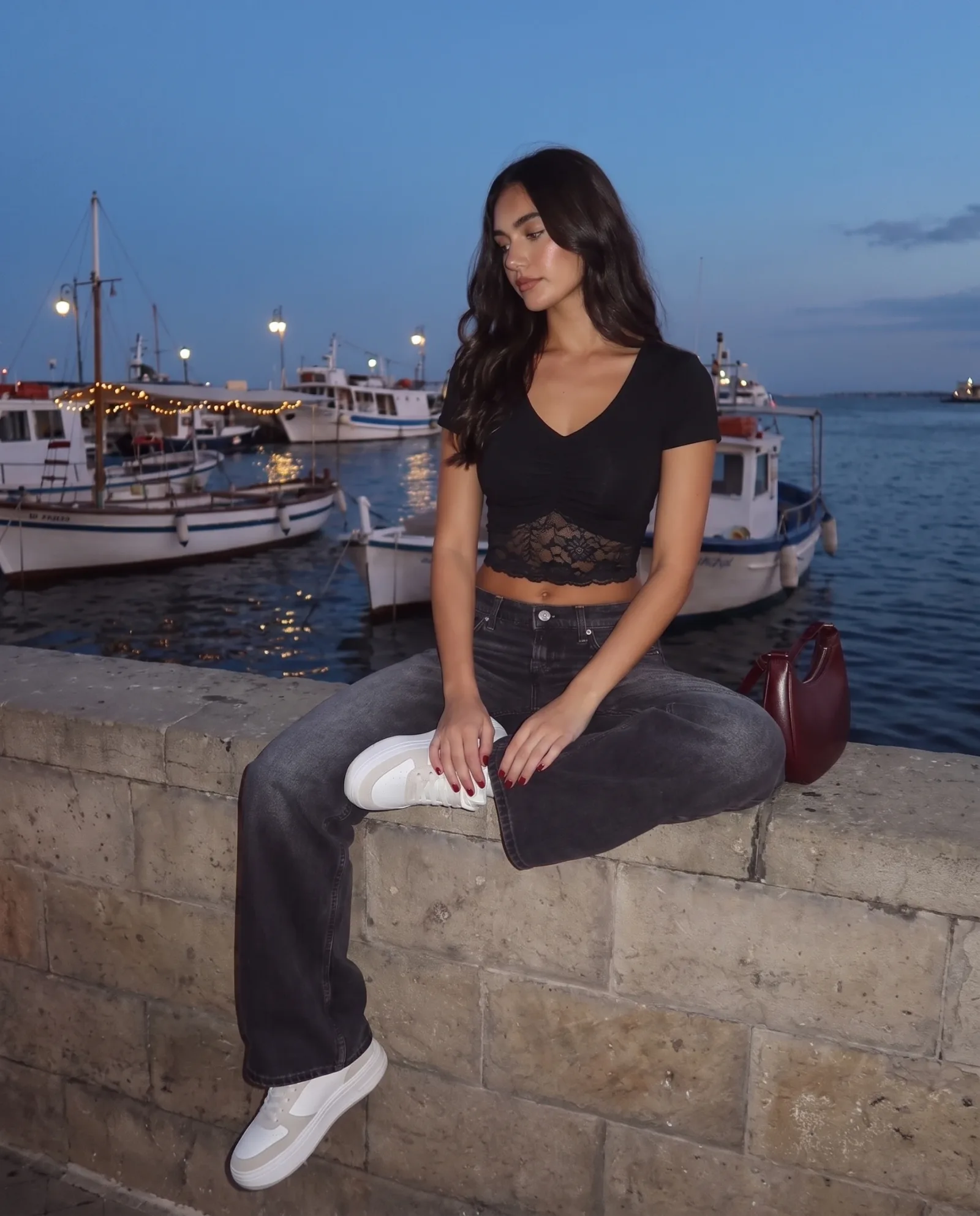 Black v-neck crop top, sitting on a harbour wall with fishing boats and string lights behind.