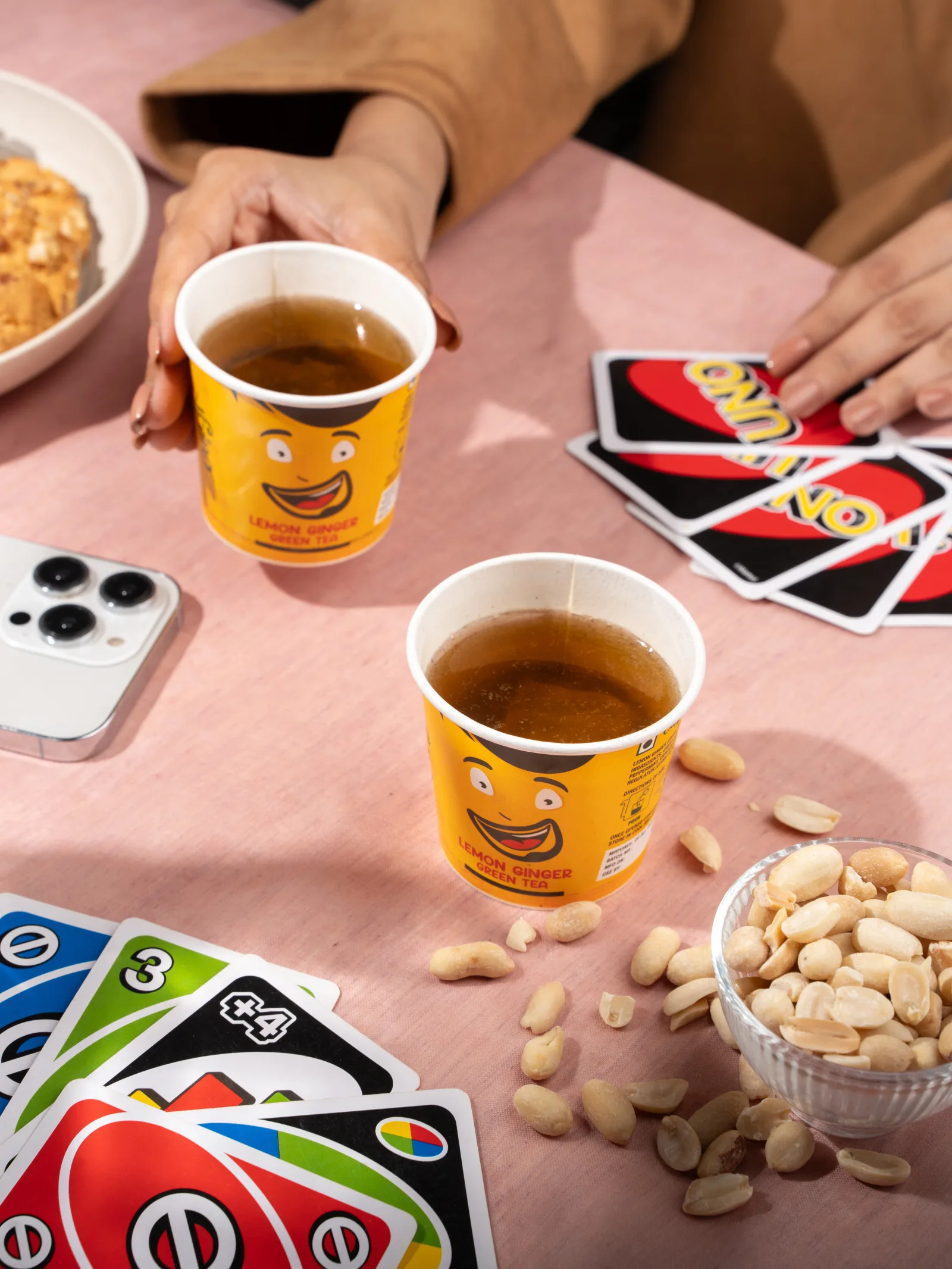 Two Lemon Ginger cups on a pink table scattered with Uno cards, peanuts and a phone, a hand reaching in.